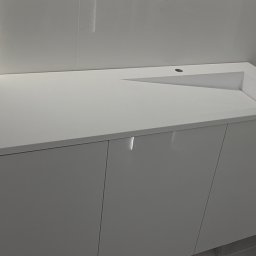 Jakub Zajac - Biała szafka łazienkowa z umywalką o geometrycznym kształcie, minimalistyczny design, gładkie fronty szafek, nowoczesne wykończenie, jasne płytki w tle.