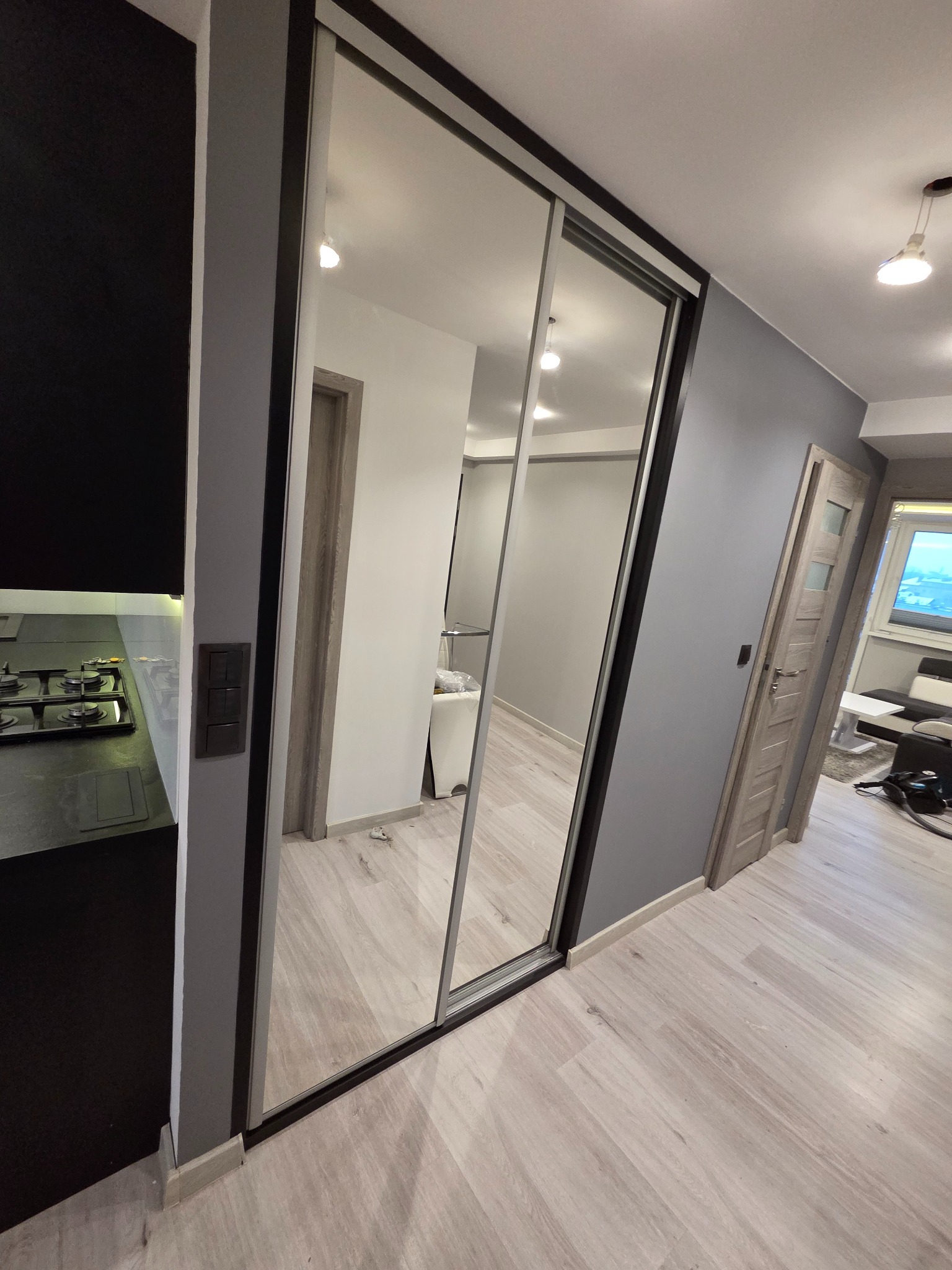Nowoczesna szafa wnękowa z lustrzanymi drzwiami przesuwnymi, widoczna w korytarzu obok czarnej zabudowy kuchennej. Minimalistyczny design, szare ściany i jasna podłoga.