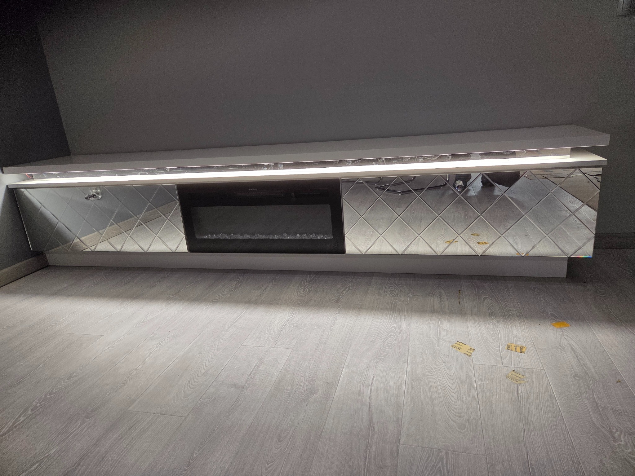 Biała szafka RTV z lustrzanymi frontami w romby i wbudowanym elektrycznym kominkiem, oświetlona listwą LED, na tle szarej ściany i podłogi.