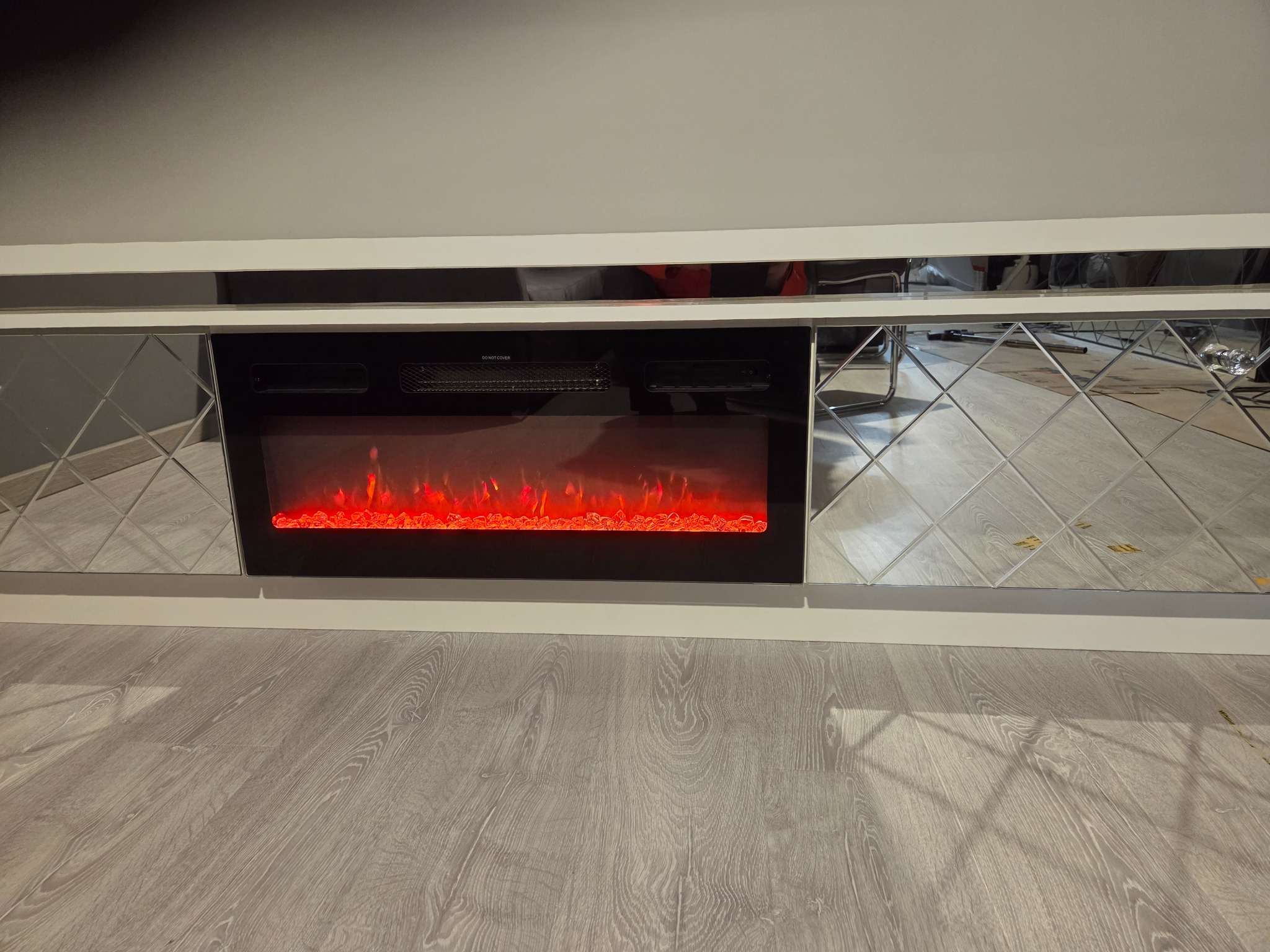 Biała szafka RTV z lustrzanymi frontami i elektrycznym kominkiem z efektem płomieni, na tle szarej ściany i podłogi z paneli. Nowoczesny design.