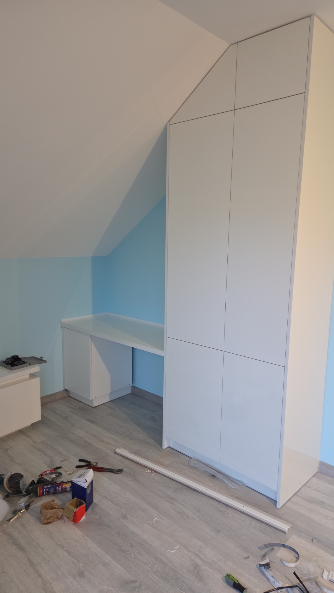 Biała szafa wnękowa z biurkiem pod skosem dachu, widoczne narzędzia na podłodze. Minimalistyczny design, jasne wnętrze, niebieska ściana w tle.