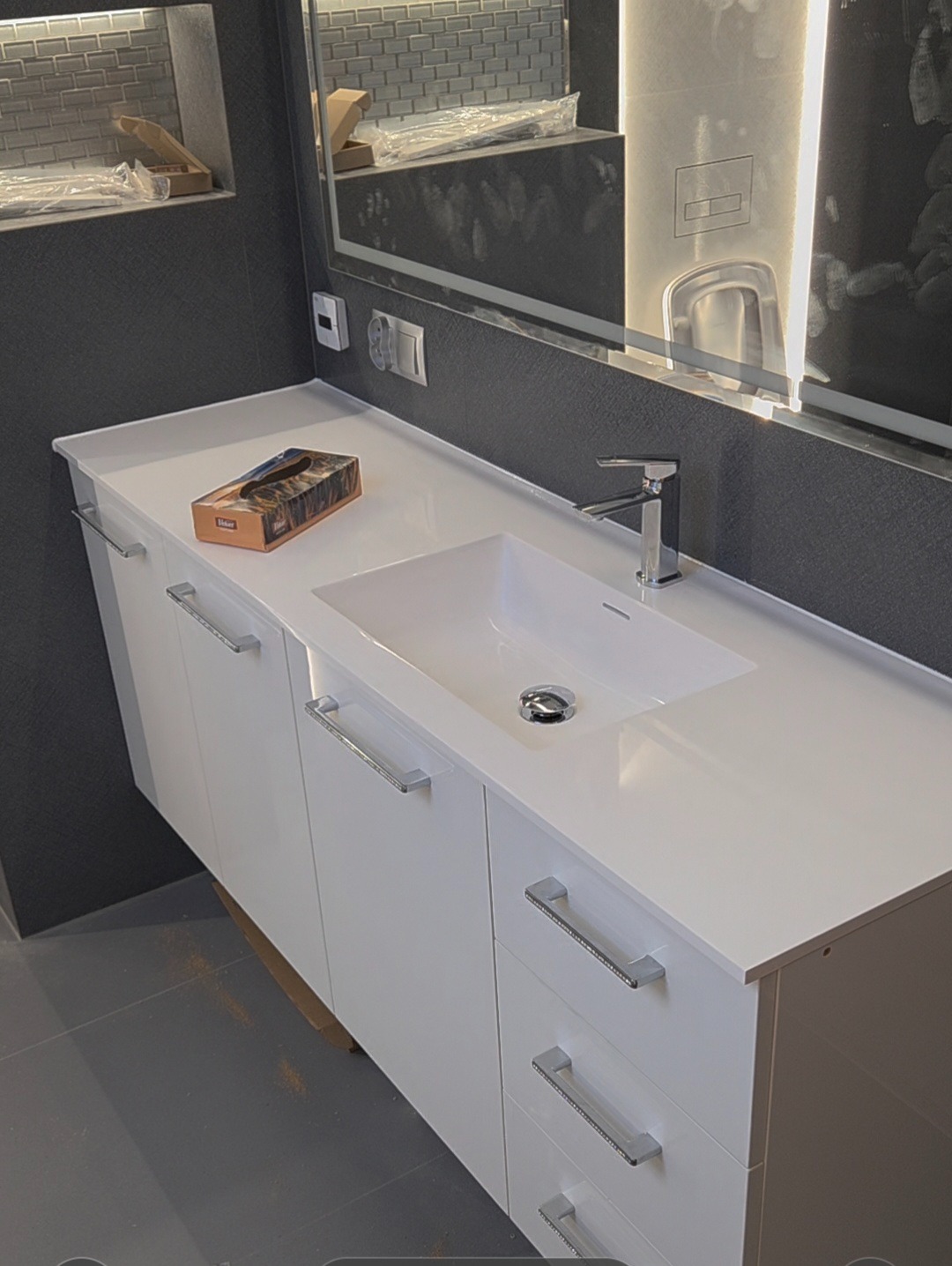 Biała szafka łazienkowa z umywalką i chromowaną armaturą, uzupełniona o pudełko chusteczek. W tle szara ściana z lustrem z podświetleniem LED.