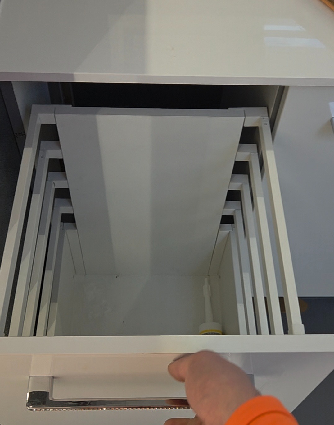 Otwarta, biała szuflada z systemem organizacji wewnętrznej. Widoczny fragment dłoni otwierającej szufladę. Minimalistyczny design, jasne oświetlenie.