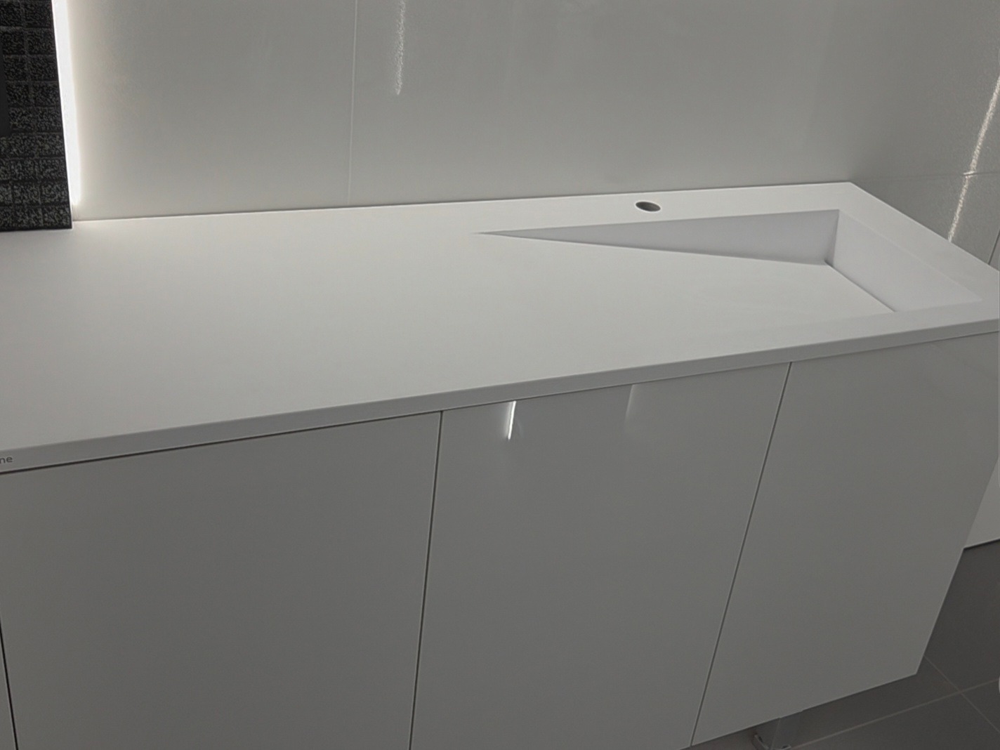 Biała szafka łazienkowa z umywalką o geometrycznym kształcie, minimalistyczny design, gładkie fronty szafek, nowoczesne wykończenie, jasne płytki w tle.