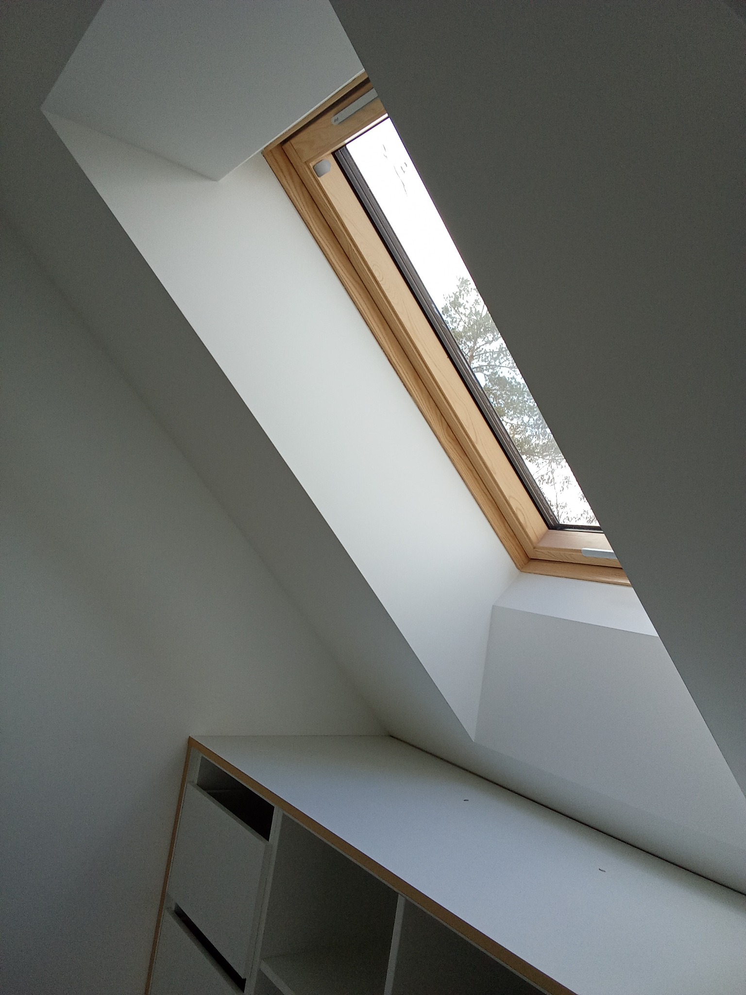 Białe poddasze ze skosem i oknem dachowym z drewnianą ramą. Widoczny fragment zabudowy meblowej z białymi frontami. Za oknem widać drzewa.