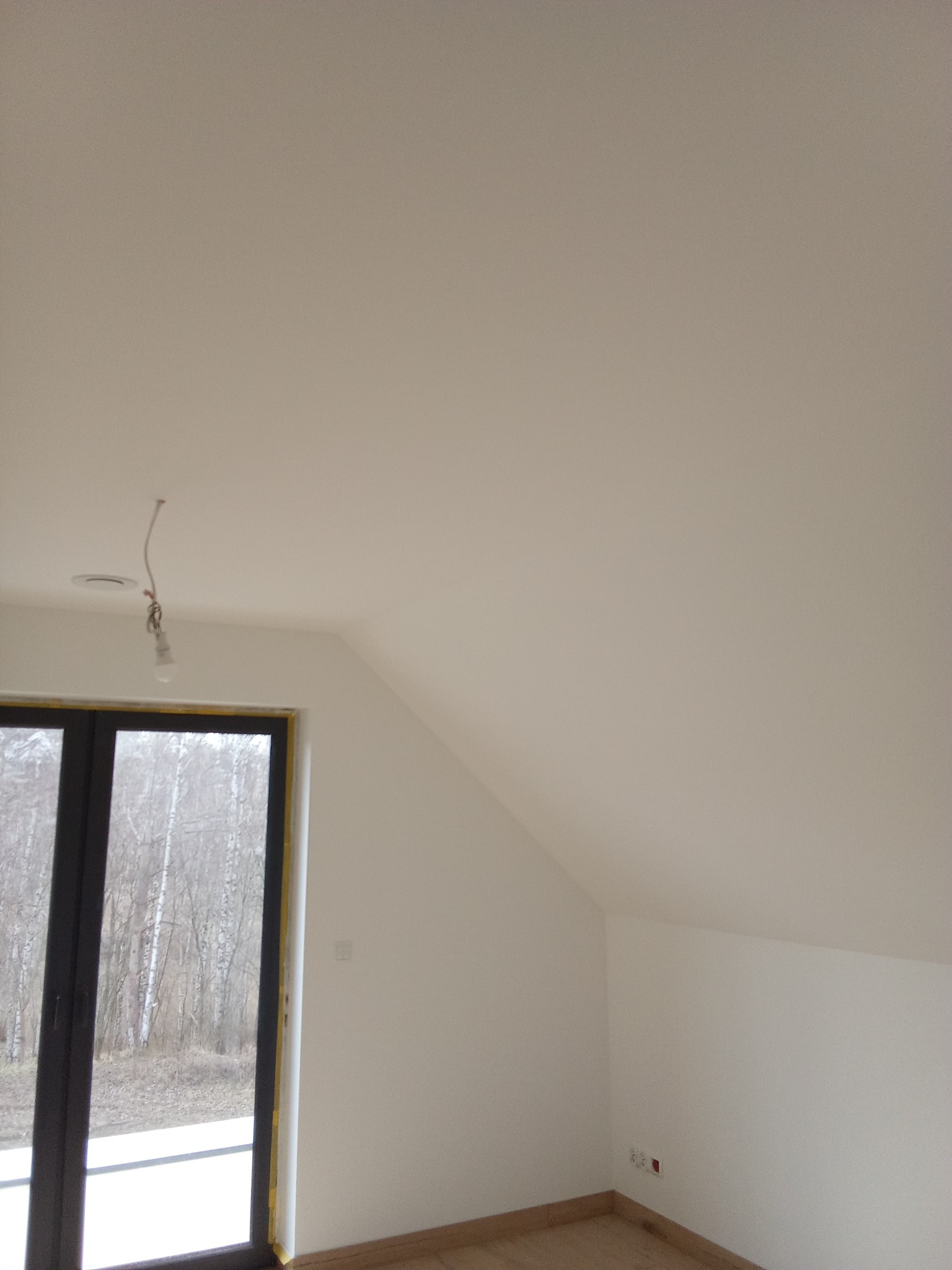 Poddasze po malowaniu na biało, widoczne okno z widokiem na las, gładkie ściany i sufit. W tle widoczne gniazdka elektryczne.