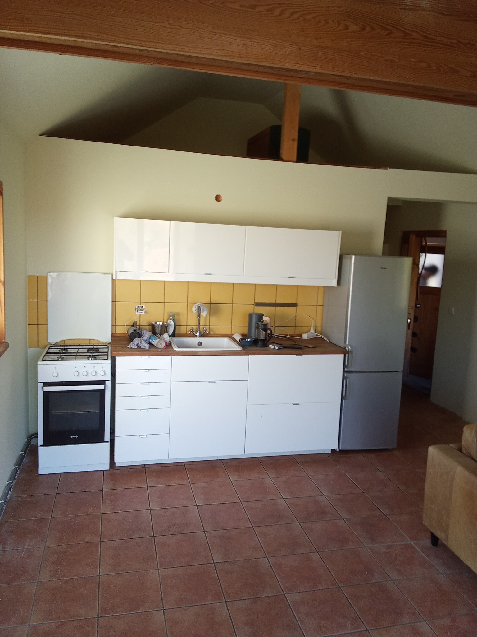 Jasna kuchnia z białymi szafkami i żółtymi płytkami nad blatem, widok na brązowe płytki podłogowe i drewniany sufit. Ukończony montaż mebli kuchennych.