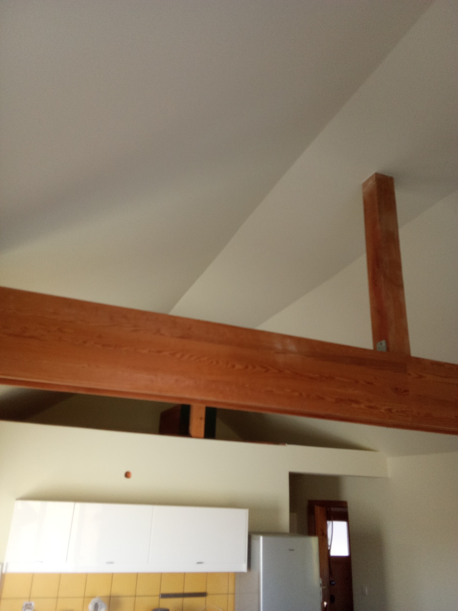 Wykończone wnętrze z drewnianą belką stropową i słupem, widok na białe szafki kuchenne i fragment lodówki. Ściany w jasnym odcieniu beżu, widoczne drzwi w tle.