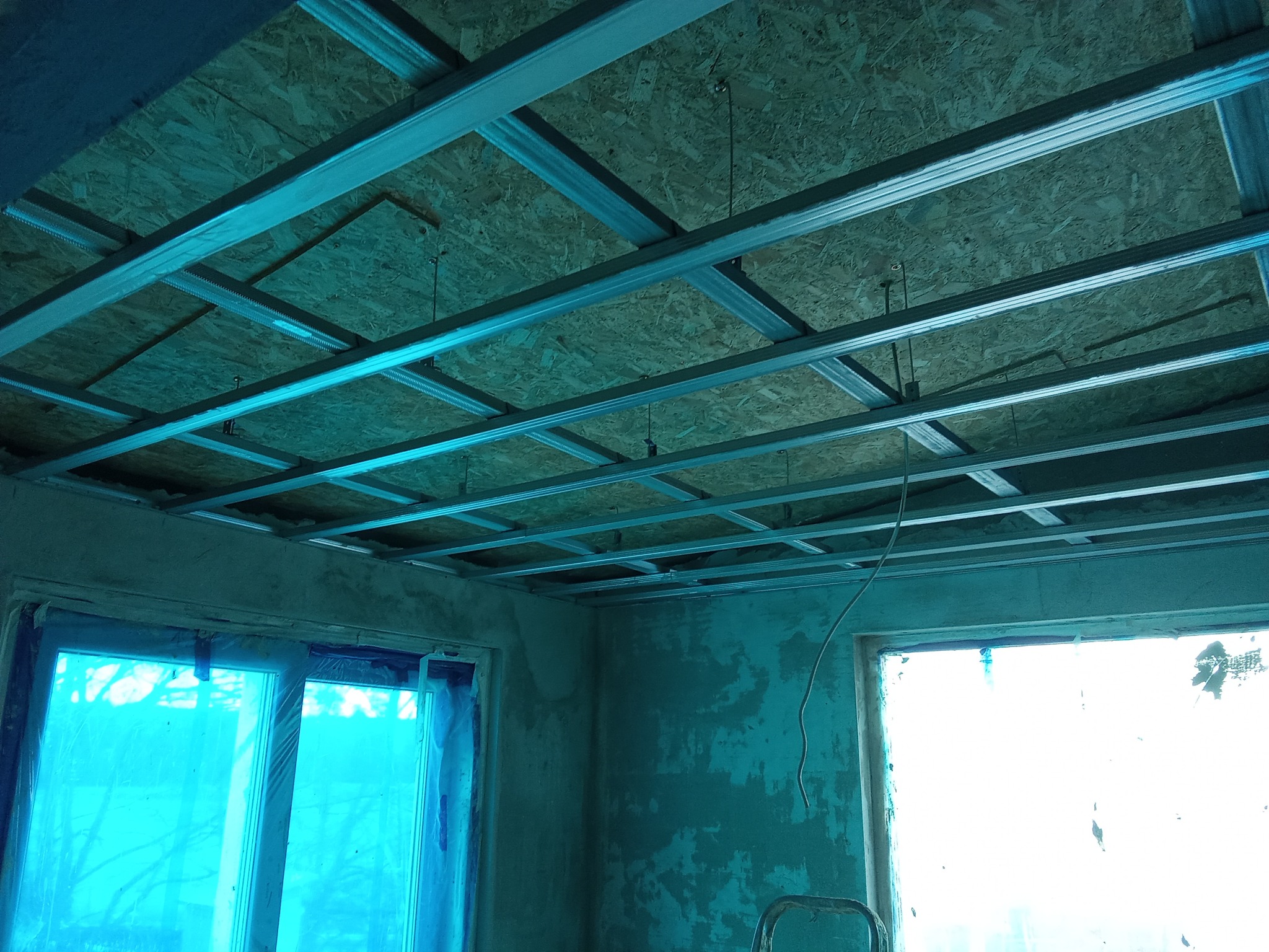 Szkielet sufitu podwieszanego z płyt karton-gips, widok z dołu. Metalowa konstrukcja kratowa, płyty OSB, okna zabezpieczone folią malarską. Wnętrze w trakcie prac wykończeniowych.