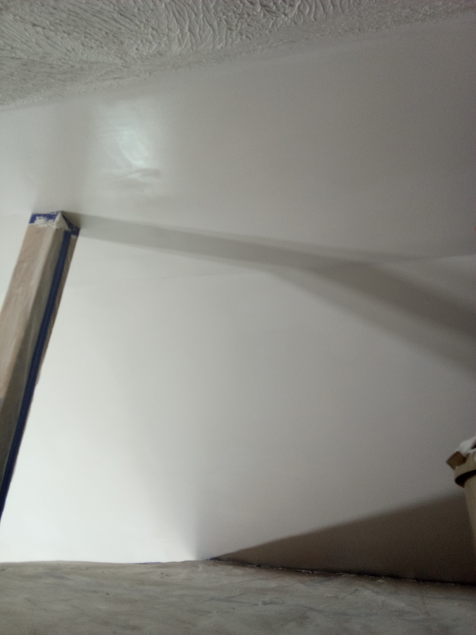 Świeżo pomalowana, biała ściana z widocznym fragmentem sufitu o nieregularnej fakturze i zabezpieczonym narożnikiem. Widoczne ślady po wałku.