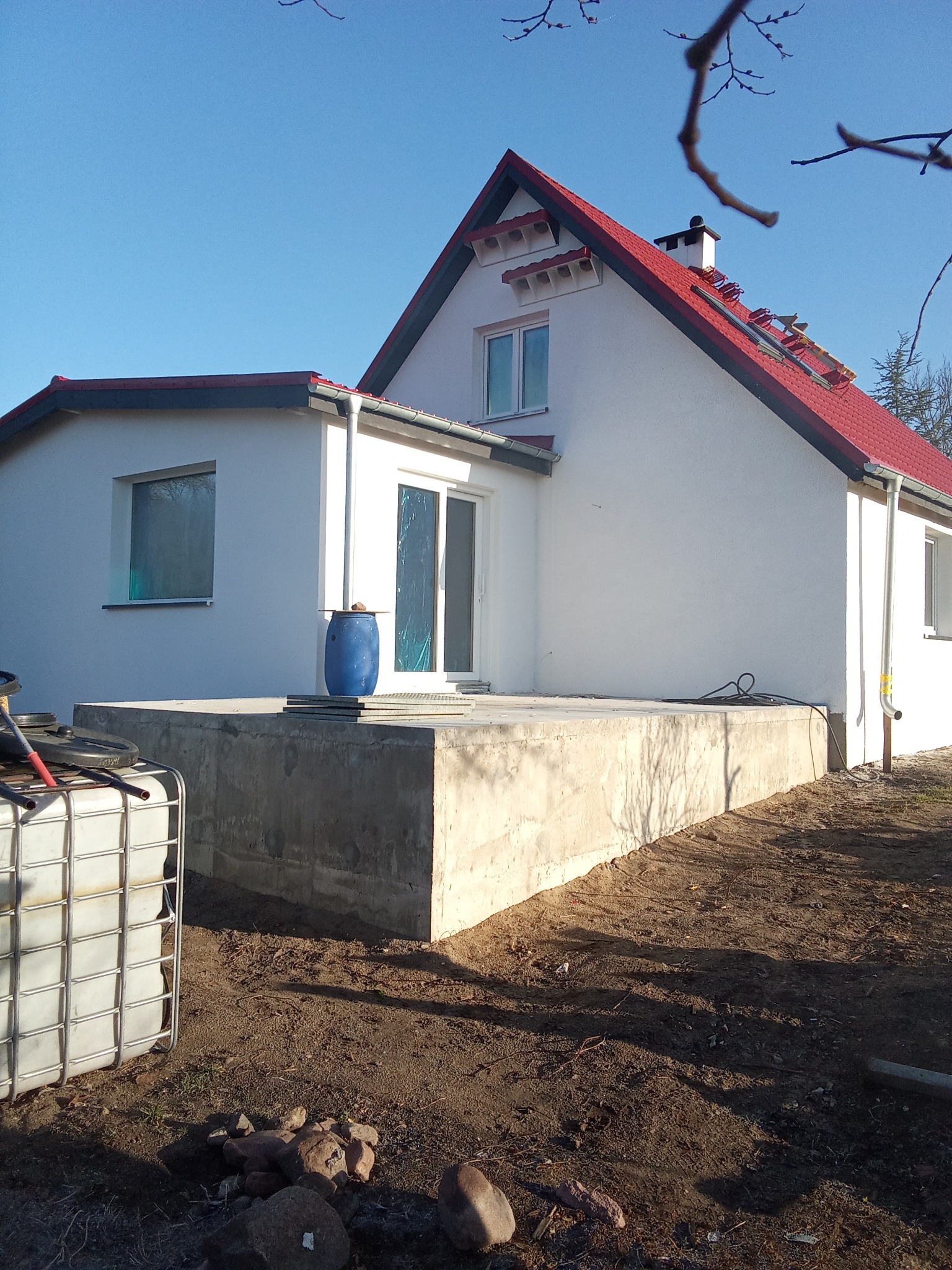 Biały dom z czerwonym dachem po remoncie, widok z zewnątrz. Częściowo widoczny fundament i elementy budowlane. Słoneczny dzień, błękitne niebo.