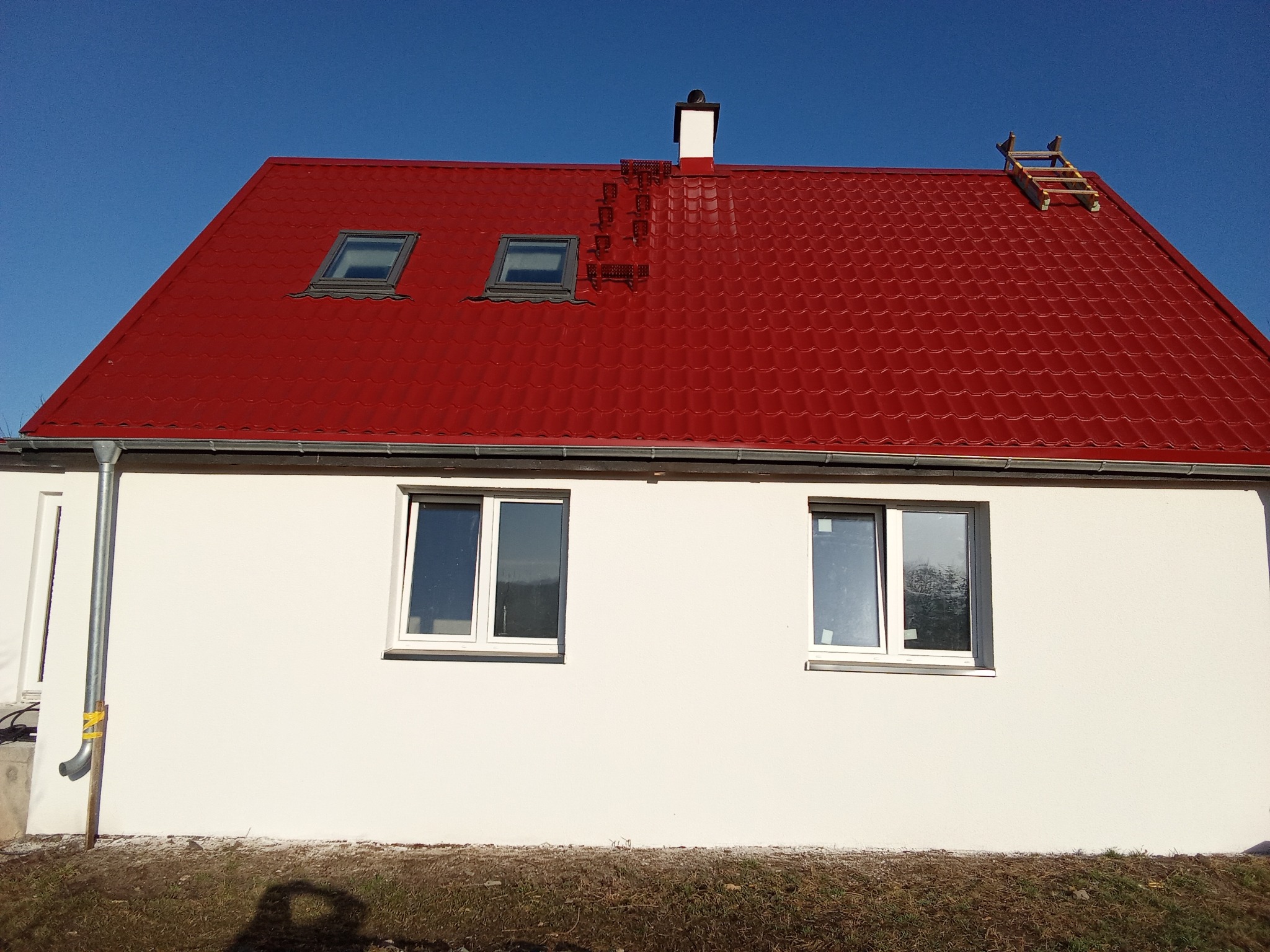 Dom z czerwoną, błyszczącą dachówką, dwoma oknami dachowymi i kominem. Na dachu drabina. Biała elewacja z dwoma oknami. Błękitne niebo w tle.