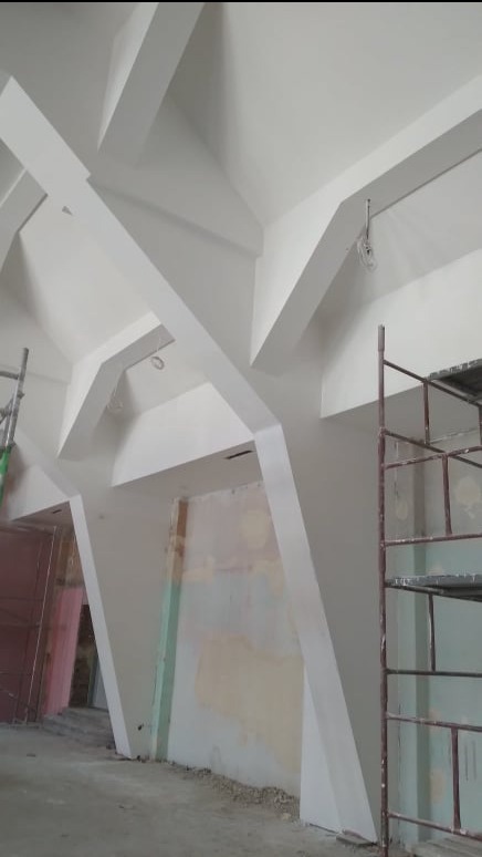 Białe, geometryczne elementy dekoracyjne sufitu i ścian w trakcie malowania. Widoczne rusztowania i ślady prac wykończeniowych. Nietypowy układ belek.