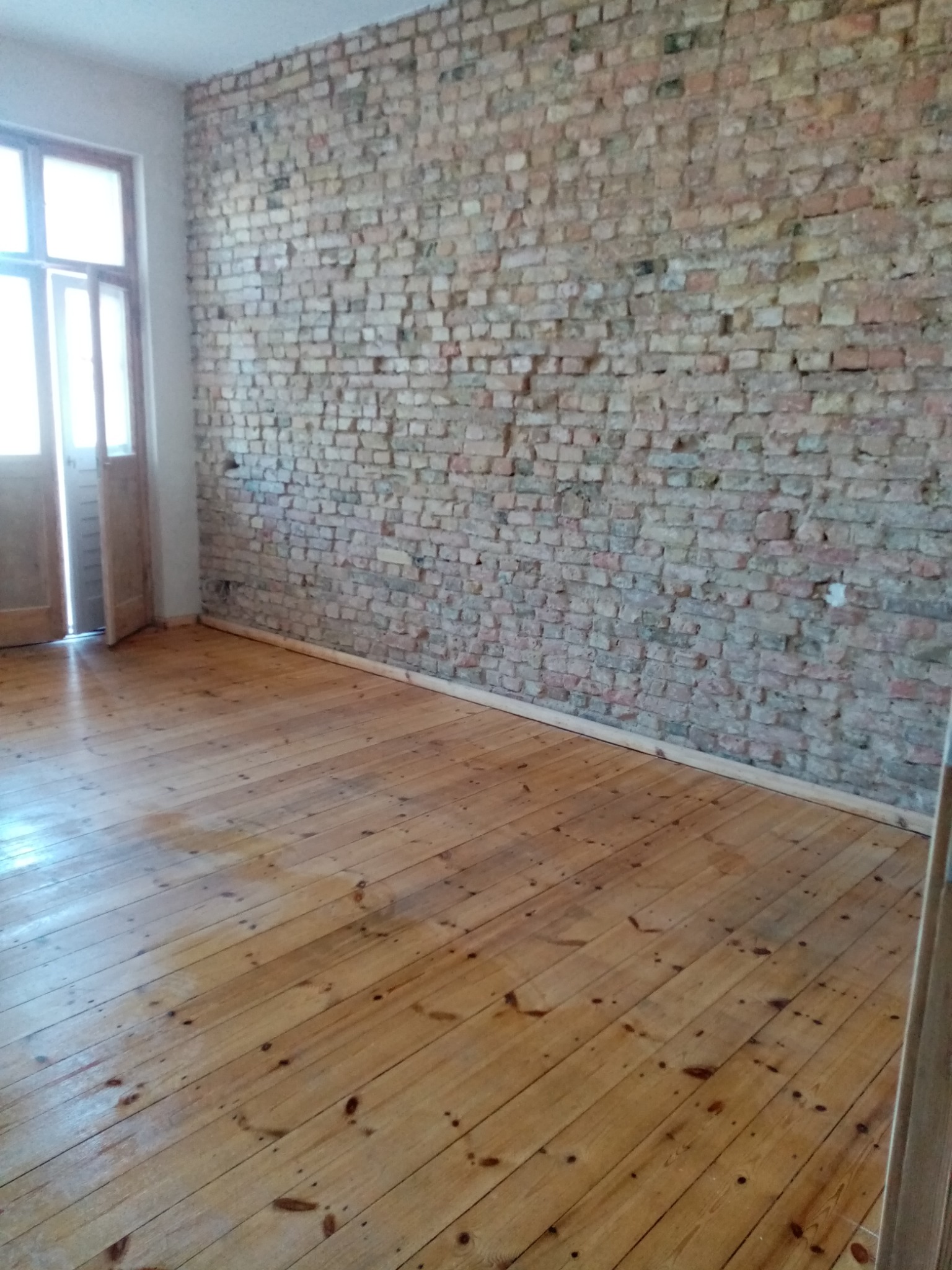 Pokój po renowacji: drewniana podłoga z widocznymi słojami i ceglana ściana z fugami. Otwarty drewniany portal drzwiowy w tle. Jasne, naturalne światło.