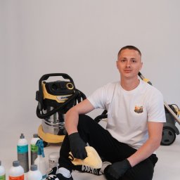 Czysty Kąt - Mężczyzna w czarnych rękawicach siedzi obok odkurzacza Karcher i środków czyszczących, trzymając ściereczkę z mikrofibry. Białe tło.