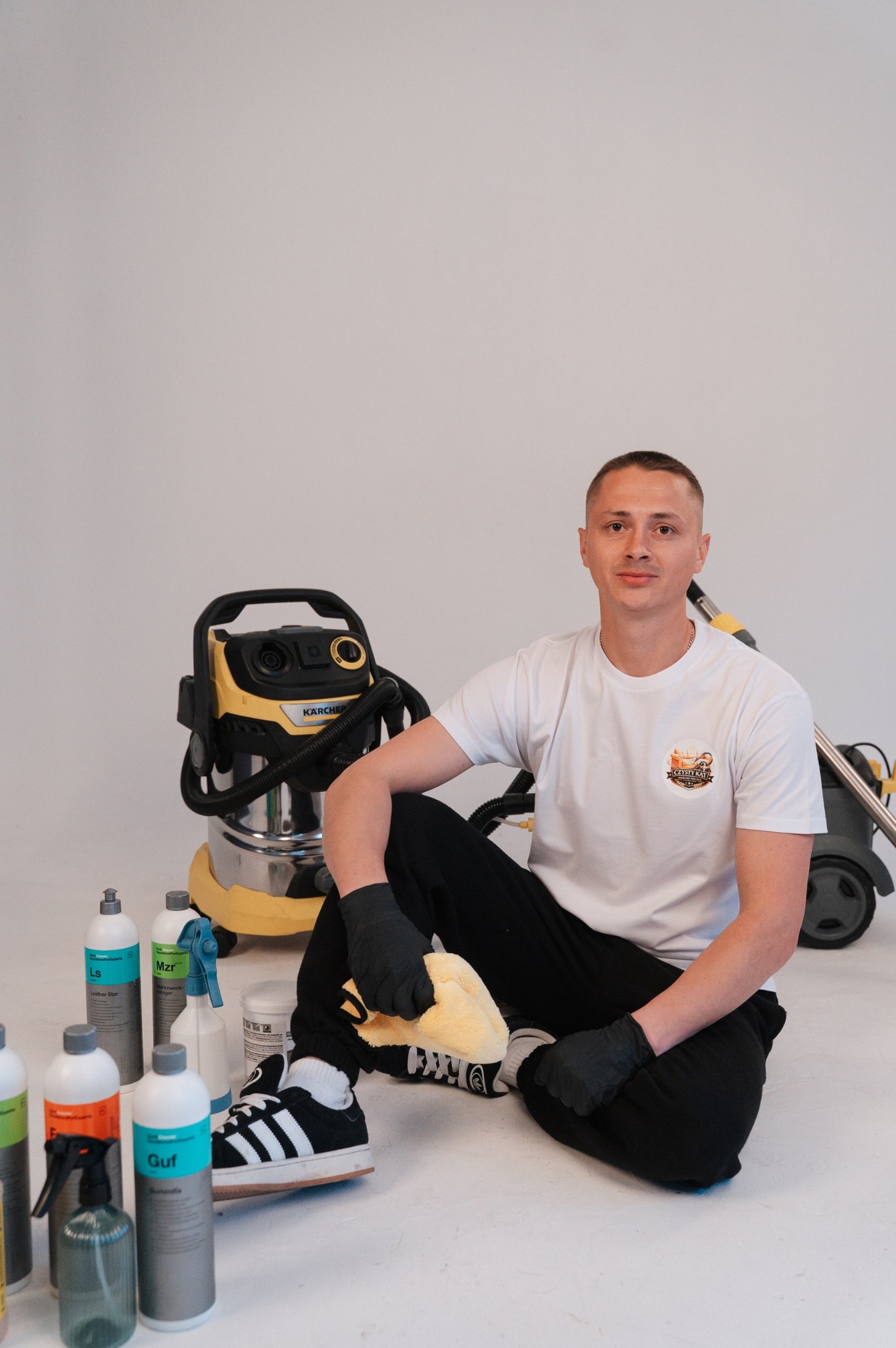 Mężczyzna w czarnych rękawicach siedzi obok odkurzacza Karcher i środków czyszczących, trzymając ściereczkę z mikrofibry. Białe tło.