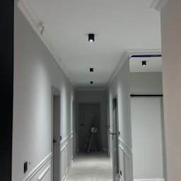 Łukasz Grzelak - Szary korytarz po remoncie z dekoracyjną sztukaterią ścienną i sufitową. Na podłodze narzędzia, w tle drabina malarska. Widok w perspektywie.