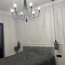 Łukasz Grzelak - Elegancka sypialnia z szarą pościelą, ozdobnymi panelami ściennymi i designerskim, czarnym żyrandolem. Minimalistyczne lampy wiszące po bokach łóżka.