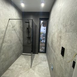 Łukasz Grzelak - Nowoczesna łazienka w szarościach z kabiną typu walk-in i oknem. Minimalistyczny design, płytki imitujące kamień, czarna armatura i oświetlenie punktowe.