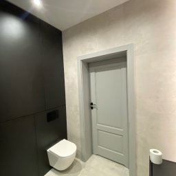 Łukasz Grzelak - Nowoczesna łazienka z podwieszaną toaletą, czarną zabudową i szarą stolarką drzwiową. Minimalistyczny design, jasne płytki imitujące beton, oświetlenie punktowe.
