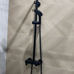 Łukasz Grzelak - Czarna bateria prysznicowa z deszczownicą na tle beżowych, wzorzystych płytek w łazience. Nowoczesny design i precyzyjny montaż.