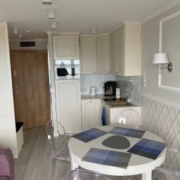 Łukasz Grzelak - Jasna kuchnia w mieszkaniu po remoncie: białe meble, okrągły stół z przezroczystymi krzesłami. Nowoczesne oświetlenie sufitowe i dekoracyjna ściana z geometrycznym wzorem.