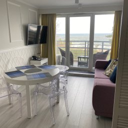 Łukasz Grzelak - Jasne wnętrze pokoju z widokiem na morze: biały stół z krzesłami, sofa z poduszkami, telewizor na ścianie z dekoracyjną fakturą. Nowoczesny design.