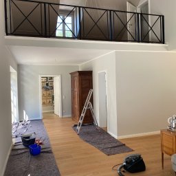 Łukasz Grzelak - Dwupoziomowe wnętrze w trakcie malowania: zabezpieczona podłoga, drabina oparta o ścianę, widoczne narzędzia malarskie. Ściany w jasnym odcieniu szarości, sufit w trakcie prac.