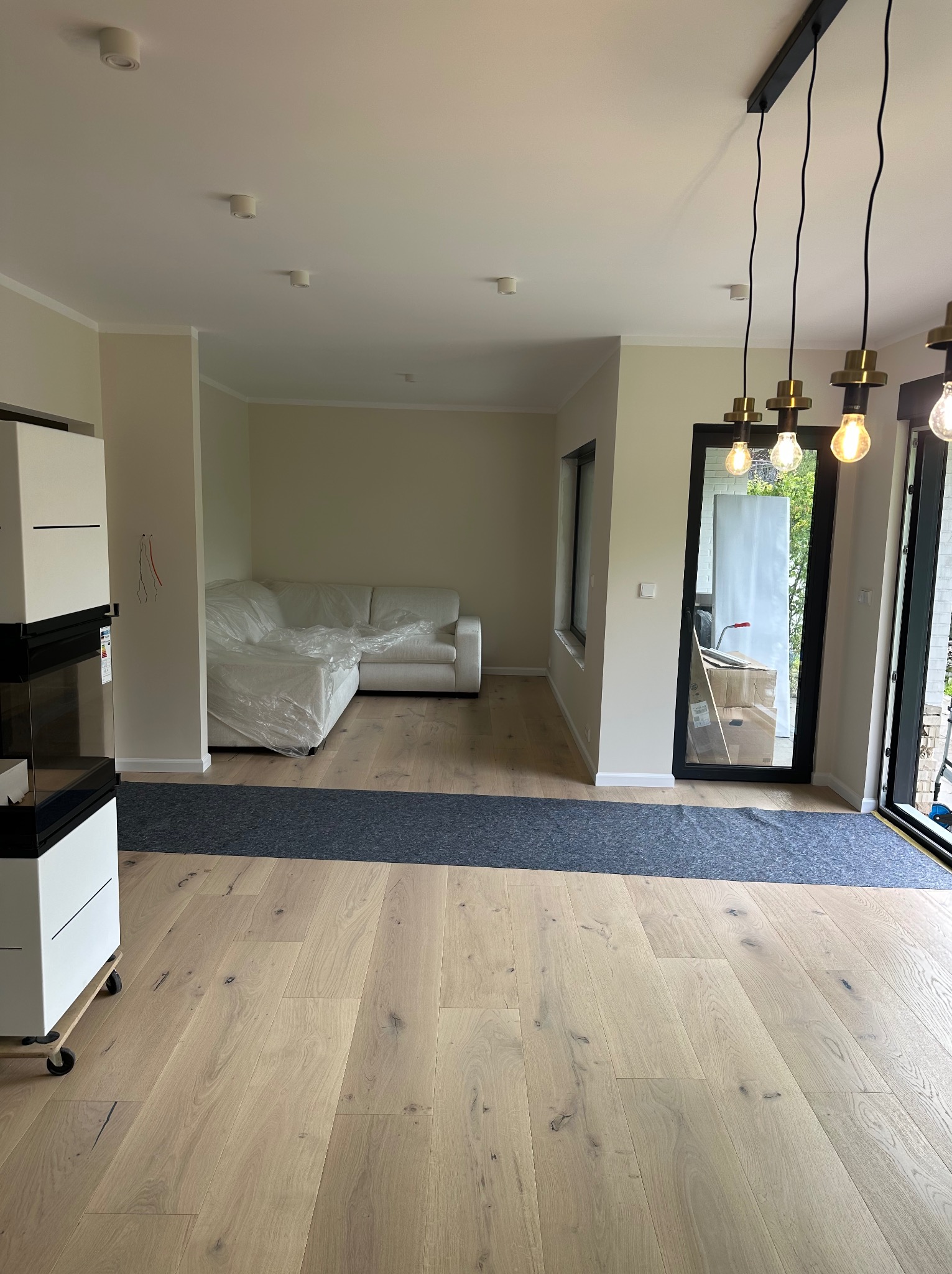Jasne drewniane panele podłogowe w nowym salonie z sofą zabezpieczoną folią. Widoczne lampy wiszące i okno balkonowe. Przygotowanie do wykończenia wnętrza.