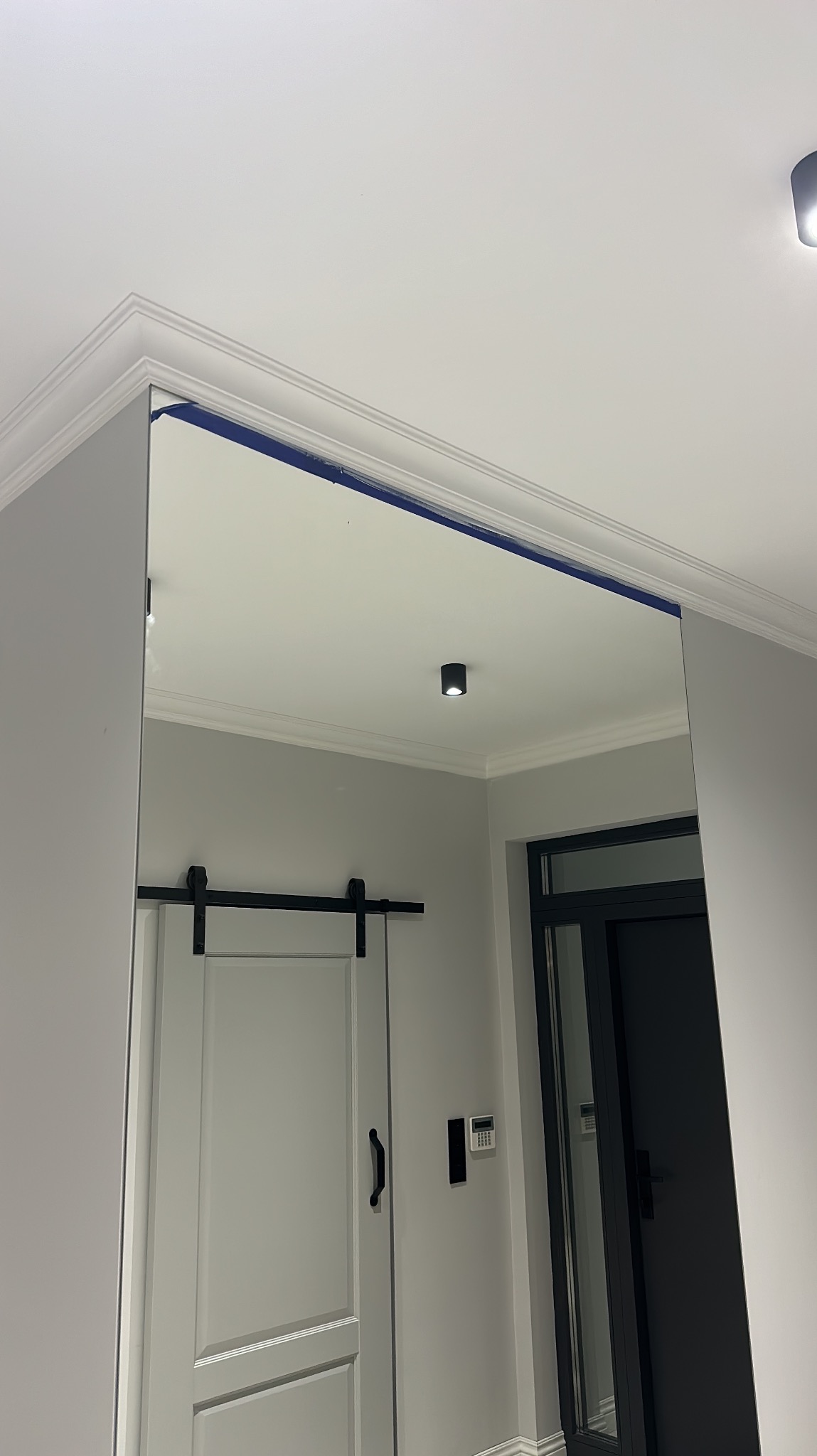 Nowoczesne wnętrze z dużym lustrem, odzwierciedlającym drzwi przesuwne w stylu stodoły i minimalistyczne oświetlenie sufitowe. Widoczne listwy dekoracyjne.