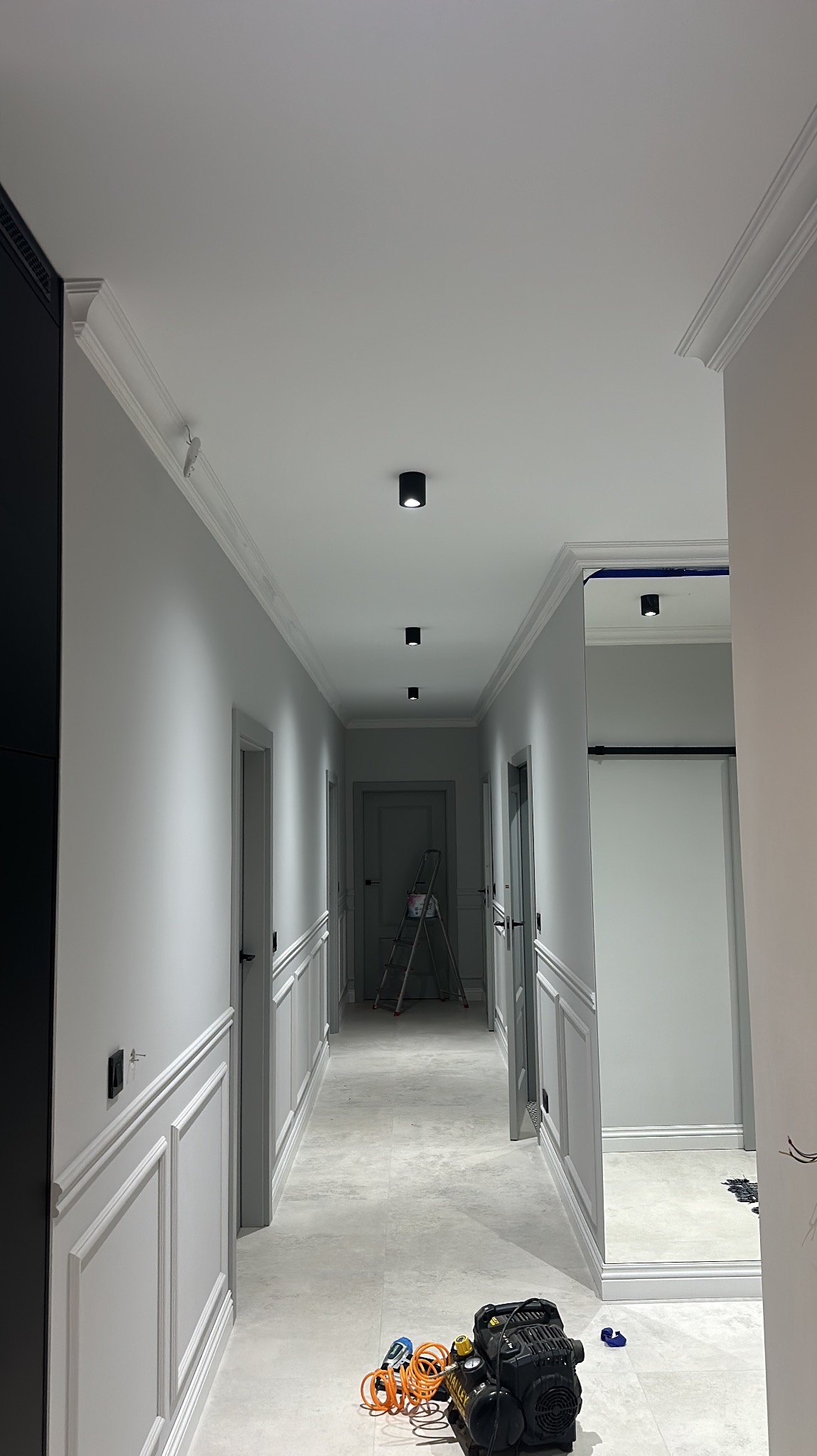 Szary korytarz po remoncie z dekoracyjną sztukaterią ścienną i sufitową. Na podłodze narzędzia, w tle drabina malarska. Widok w perspektywie.