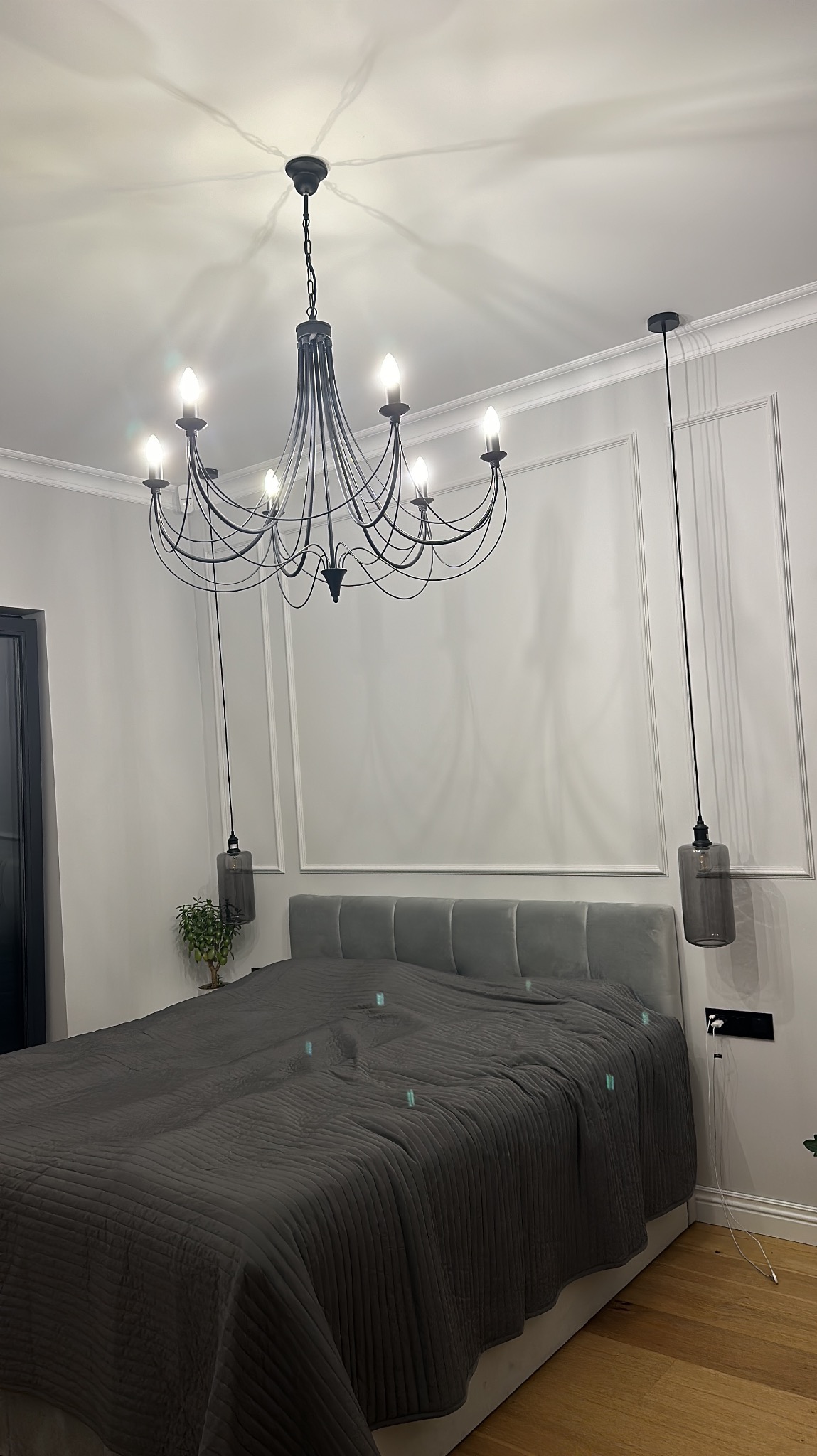 Elegancka sypialnia z szarą pościelą, ozdobnymi panelami ściennymi i designerskim, czarnym żyrandolem. Minimalistyczne lampy wiszące po bokach łóżka.