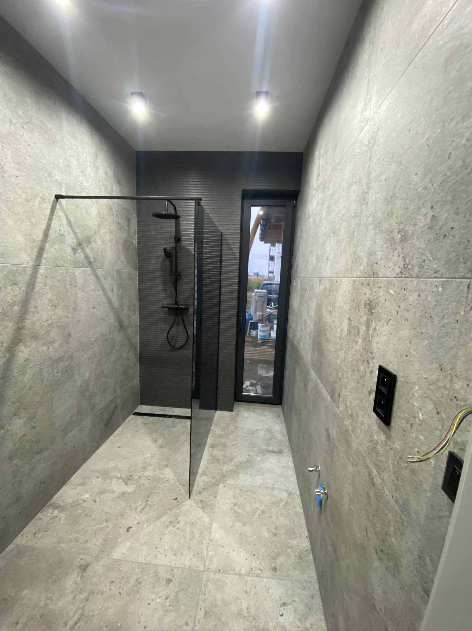 Nowoczesna łazienka w szarościach z kabiną typu walk-in i oknem. Minimalistyczny design, płytki imitujące kamień, czarna armatura i oświetlenie punktowe.