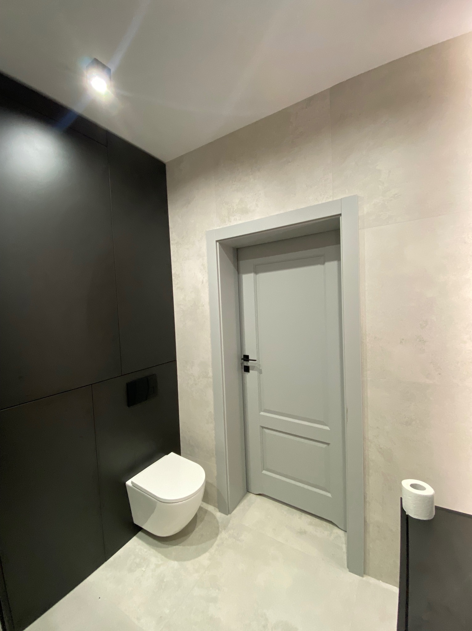 Nowoczesna łazienka z podwieszaną toaletą, czarną zabudową i szarą stolarką drzwiową. Minimalistyczny design, jasne płytki imitujące beton, oświetlenie punktowe.