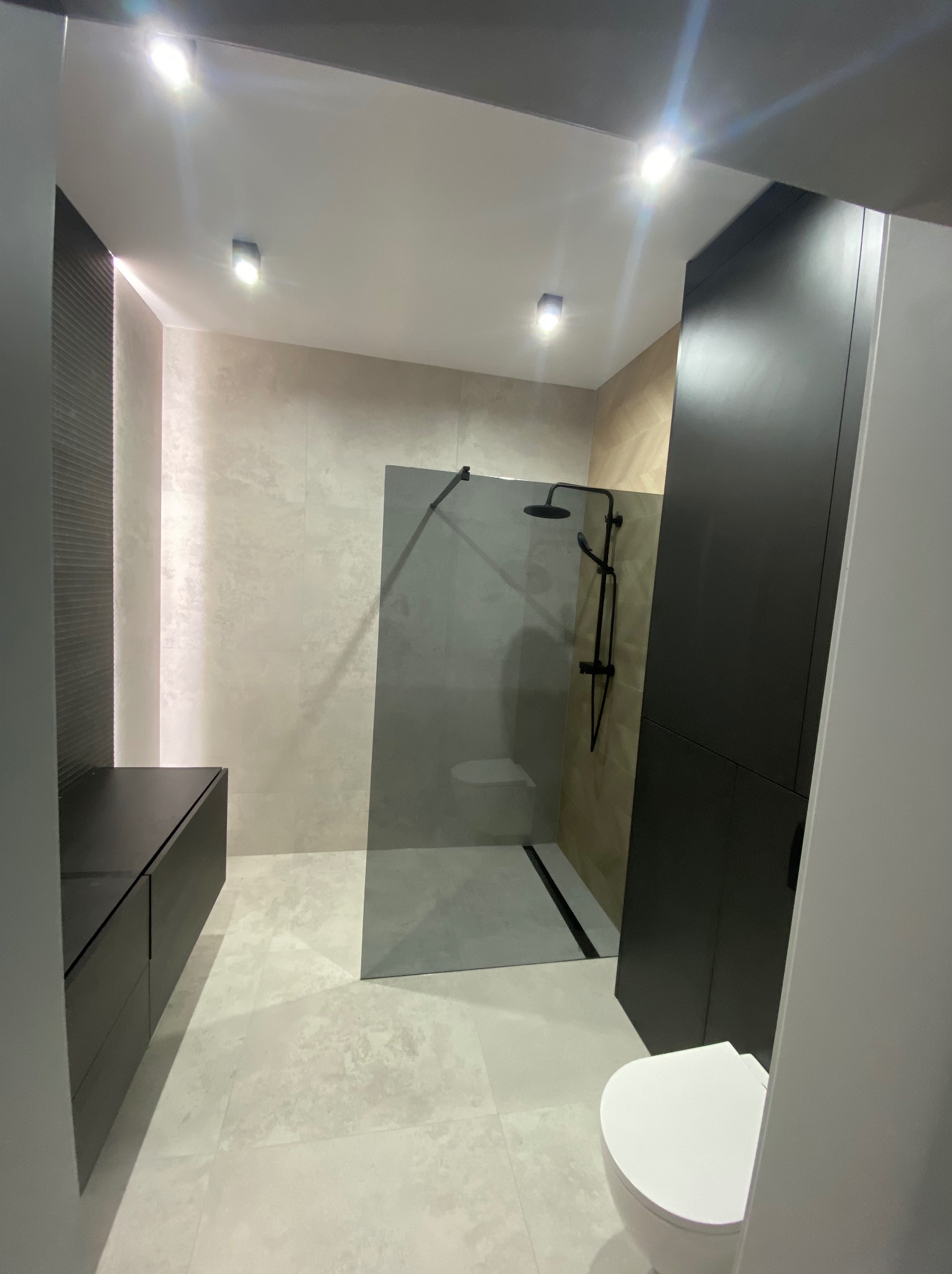 Nowoczesna łazienka z kabiną typu walk-in, czarną armaturą i szafkami w kolorze antracytu. Jasne płytki podłogowe i ścienne tworzą spójną całość.