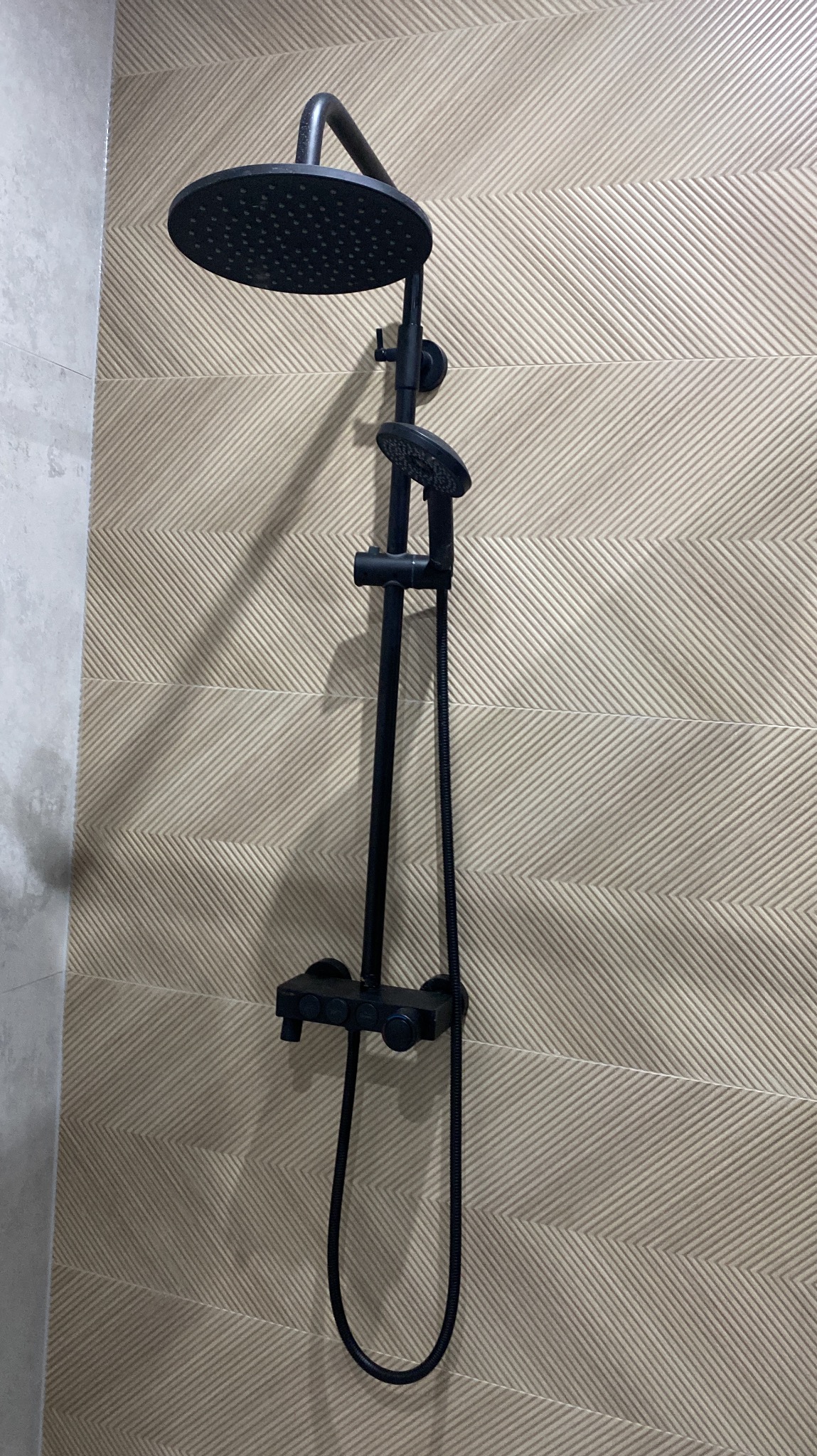 Czarna bateria prysznicowa z deszczownicą na tle beżowych, wzorzystych płytek w łazience. Nowoczesny design i precyzyjny montaż.