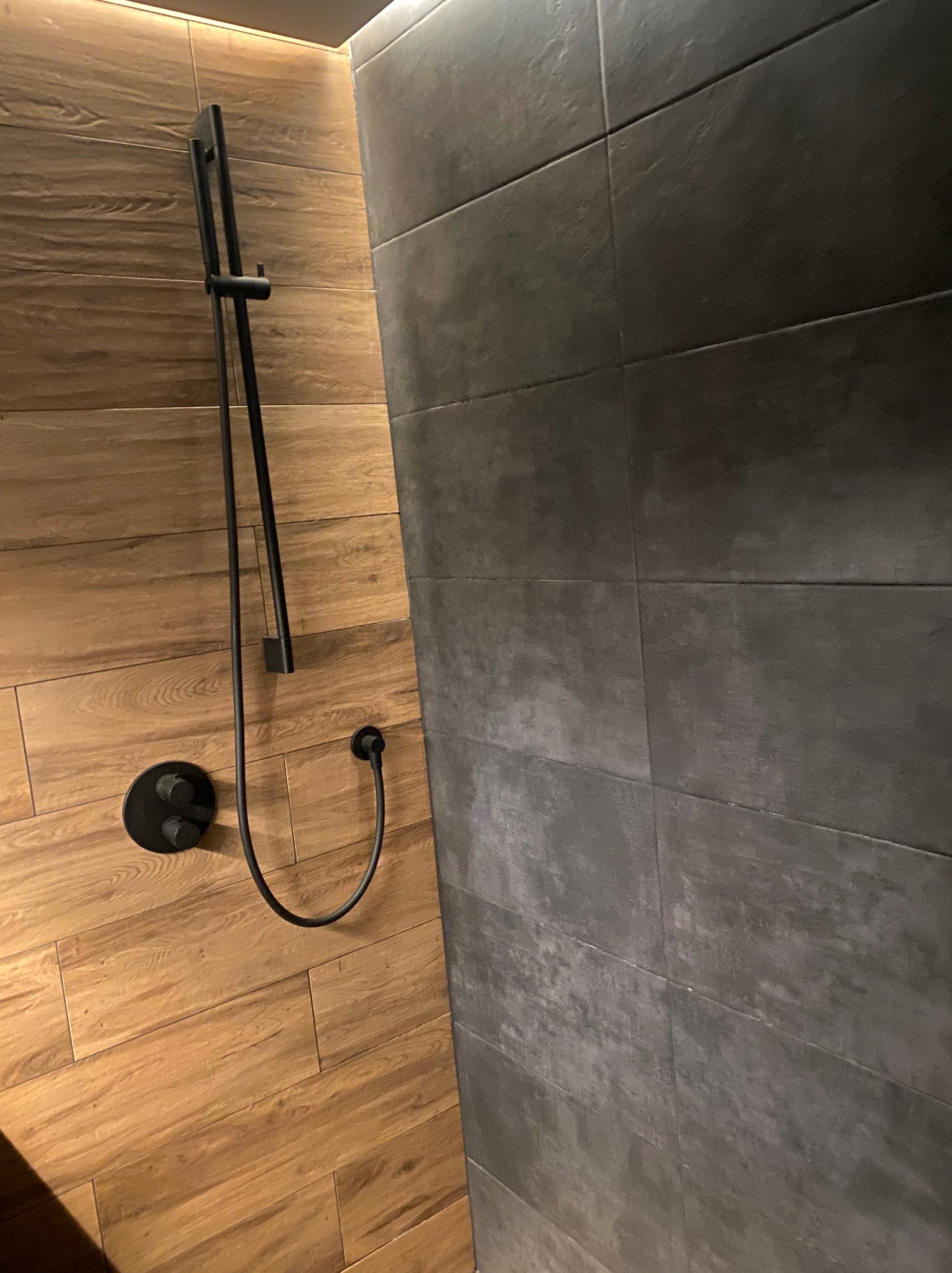 Nowoczesna kabina prysznicowa z drewnopodobnymi płytkami i czarną armaturą, minimalistyczny design, oświetlenie LED w górnej części kadru.