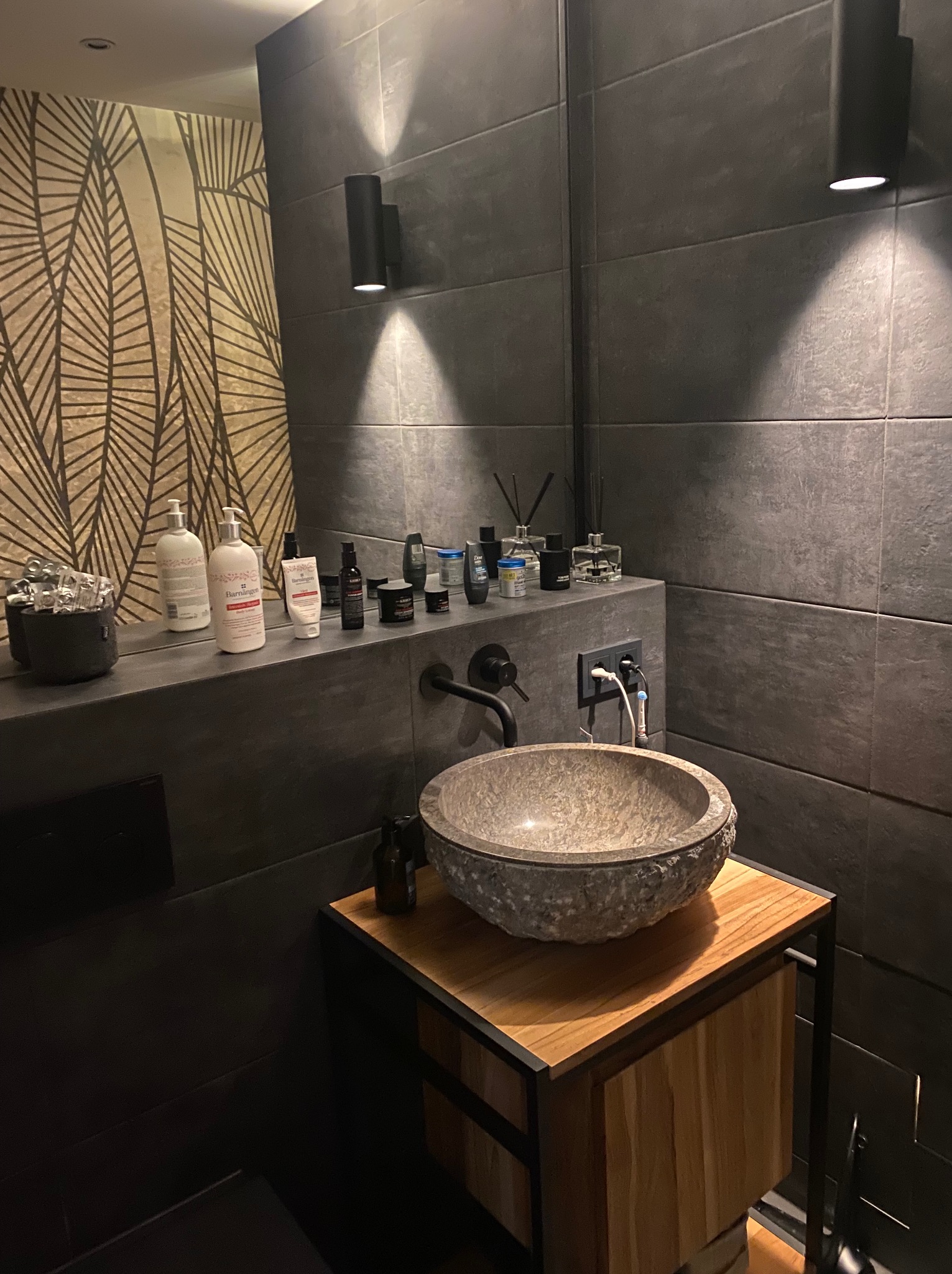 Nowoczesna łazienka z kamienną umywalką na drewnianej szafce, ciemne płytki na ścianach i designerskie oświetlenie. Uwagę przykuwa wzorzysta tapeta odbijająca się w lustrze.