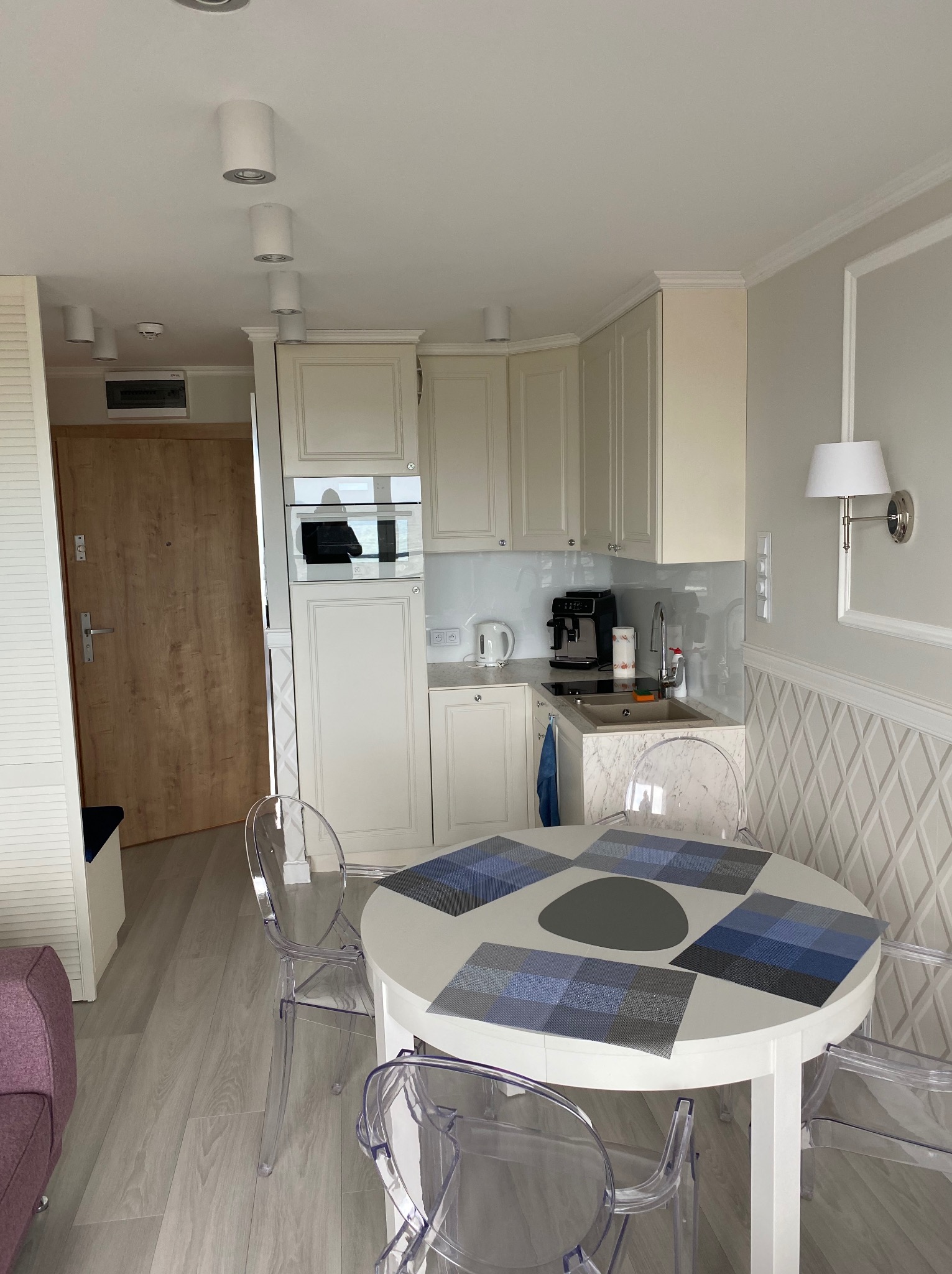 Jasna kuchnia w mieszkaniu po remoncie: białe meble, okrągły stół z przezroczystymi krzesłami. Nowoczesne oświetlenie sufitowe i dekoracyjna ściana z geometrycznym wzorem.