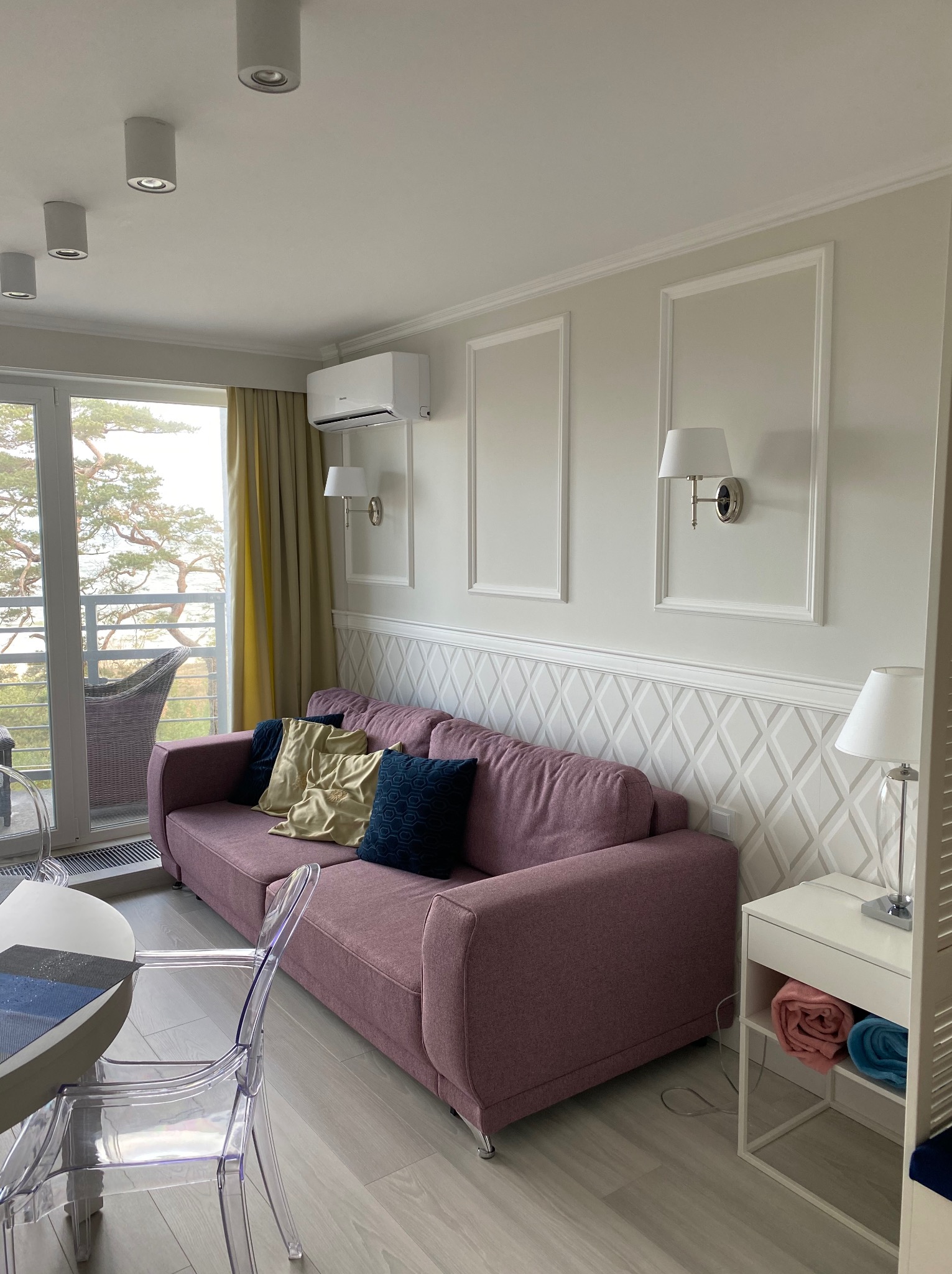 Jasne wnętrze salonu z balkonem: różowa sofa z poduszkami, designerskie lampy ścienne, wzorzysta tapeta i przezroczyste krzesła przy okrągłym stole.