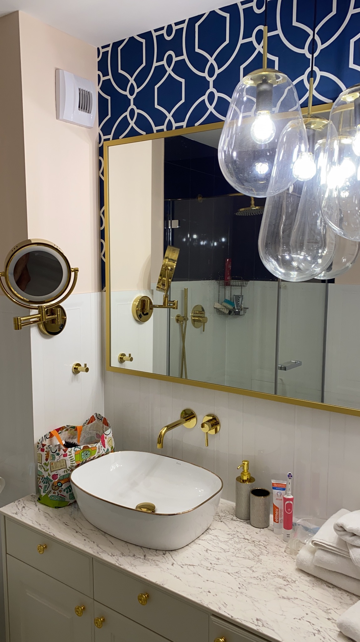 Elegancka łazienka z umywalką nablatową, złotą armaturą i lustrem w złotej ramie. Na ścianie wzorzysta tapeta w odcieniach bieli i granatu. Widoczne oświetlenie w stylu glamour.