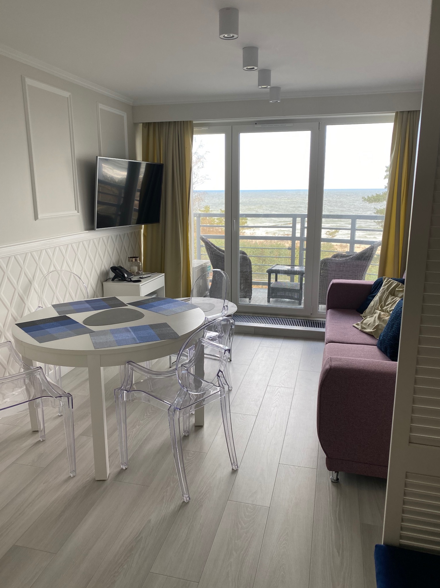 Jasne wnętrze pokoju z widokiem na morze: biały stół z krzesłami, sofa z poduszkami, telewizor na ścianie z dekoracyjną fakturą. Nowoczesny design.