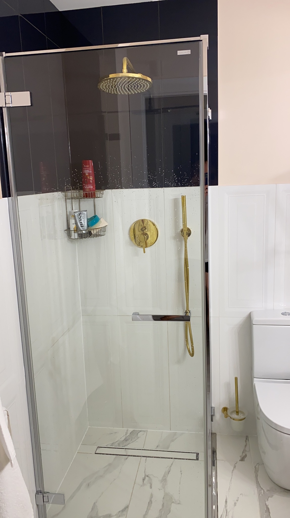 Nowoczesna kabina prysznicowa z deszczownicą w kolorze złotym, białe płytki imitujące panele, marmurowa podłoga i czarne akcenty na górze kabiny.