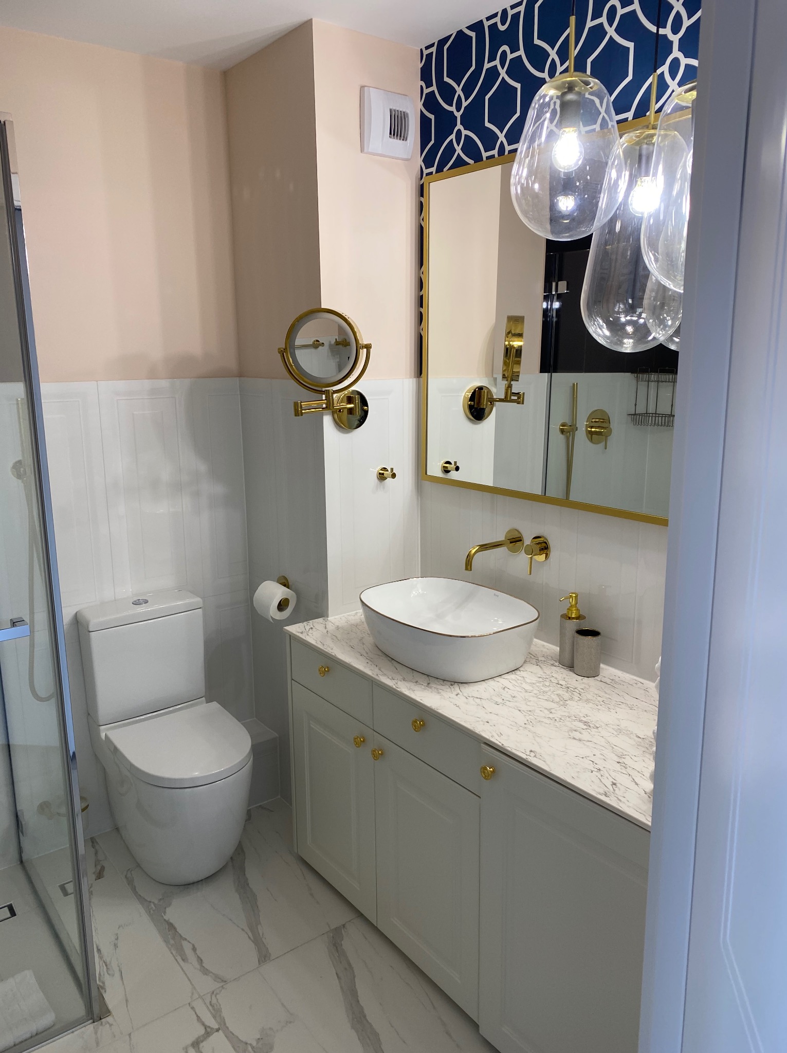 Elegancka łazienka po metamorfozie: białe płytki, marmurowa podłoga, złote akcenty armatury i oświetlenia, umywalka nablatowa i kabina prysznicowa.