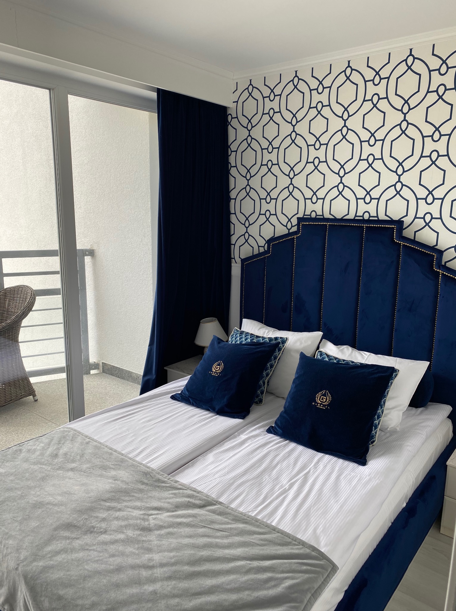 Elegancka sypialnia z balkonem: granatowe wezgłowie łóżka, wzorzysta tapeta, zasłony w odcieniu granatu i szary pled. Widok na balkon z krzesłem z rattanu.