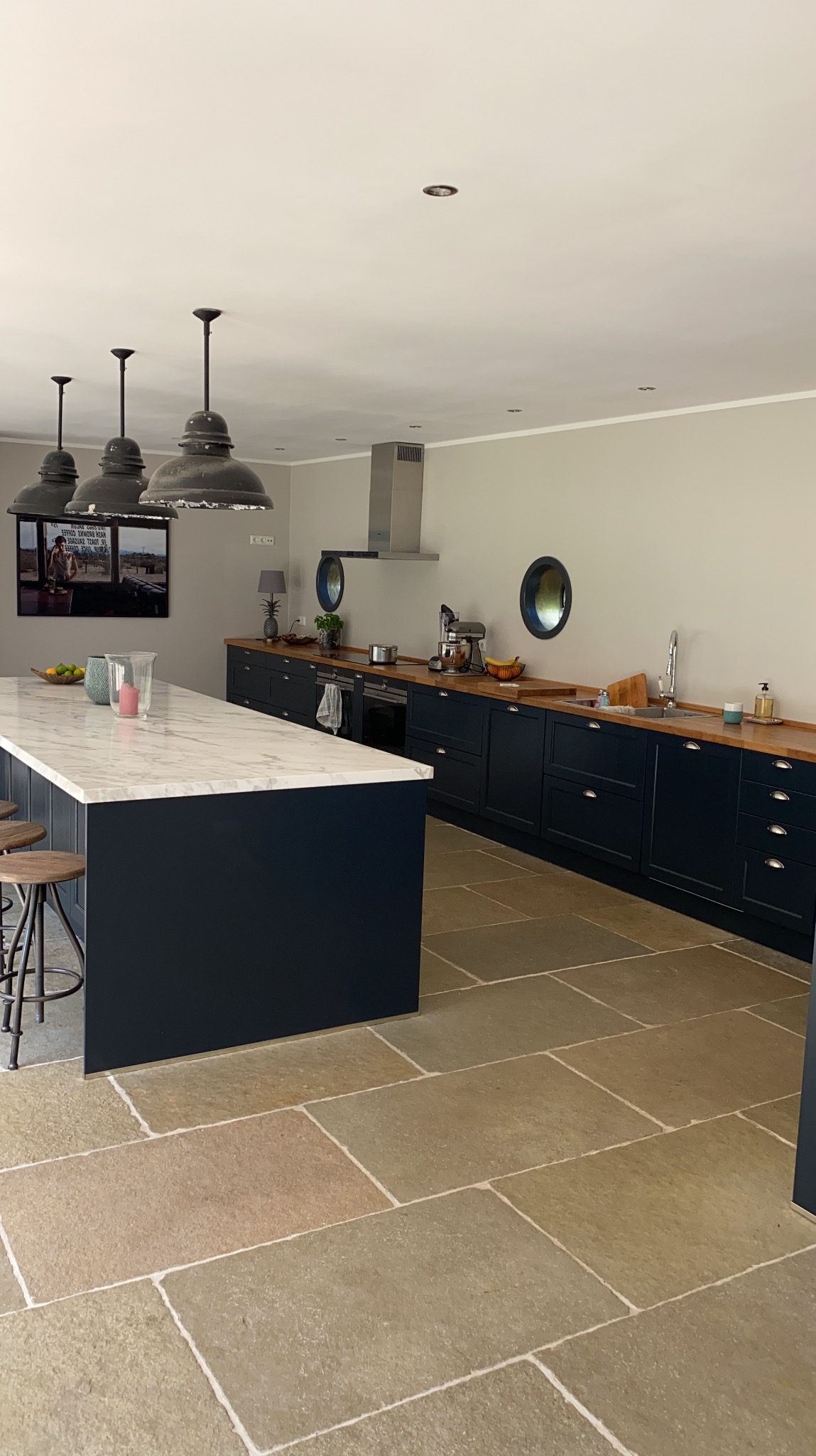 Nowoczesna kuchnia z ciemnymi szafkami, drewnianym blatem i wyspą z marmurowym blatem. Kamienna podłoga i okrągłe okna dodają unikalnego charakteru.