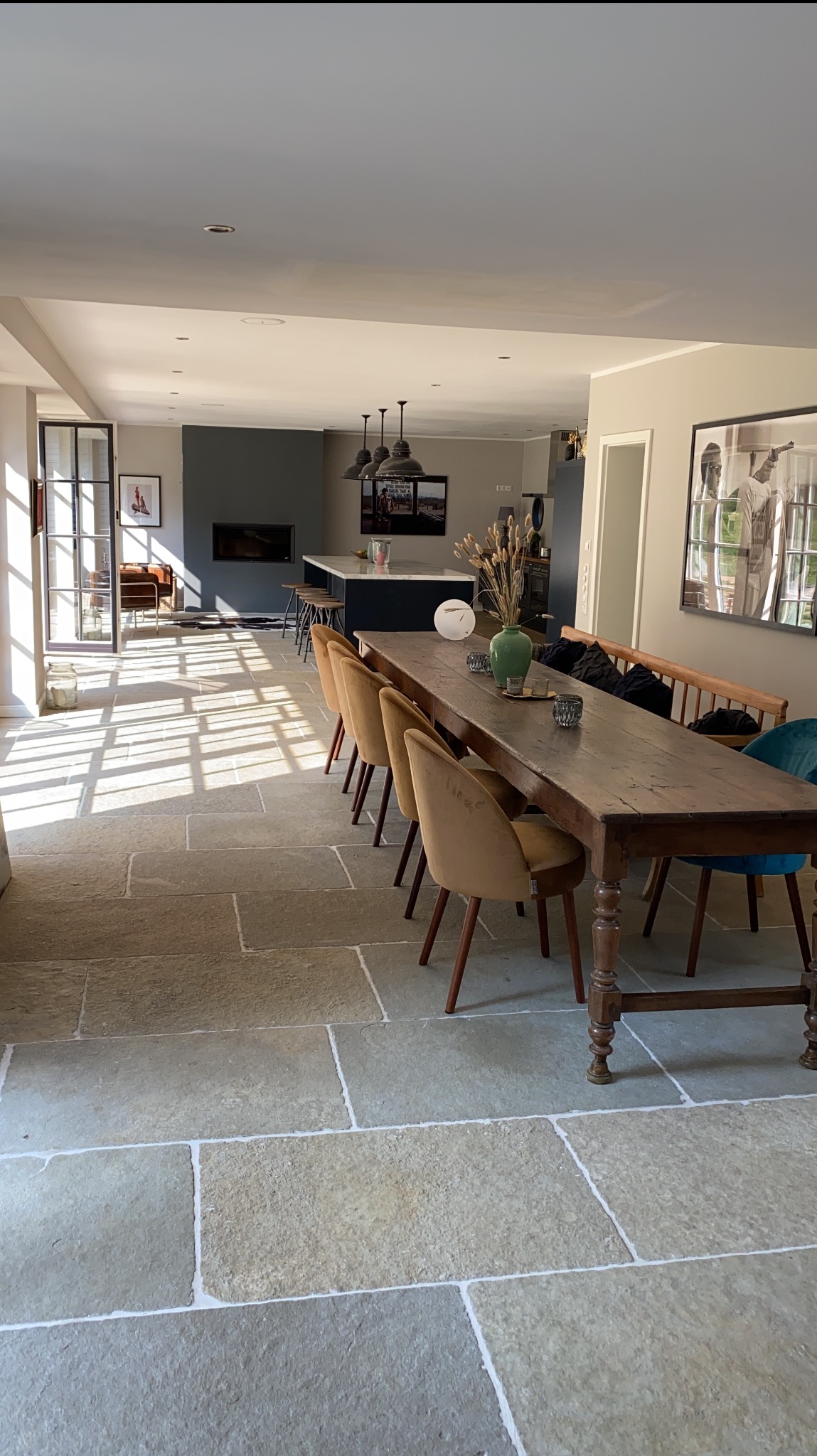 Jasne, przestronne wnętrze z kamienną podłogą i długim drewnianym stołem. W tle widoczna kuchnia z wyspą i nowoczesnym oświetleniem. Promienie słoneczne wpadają przez okno.