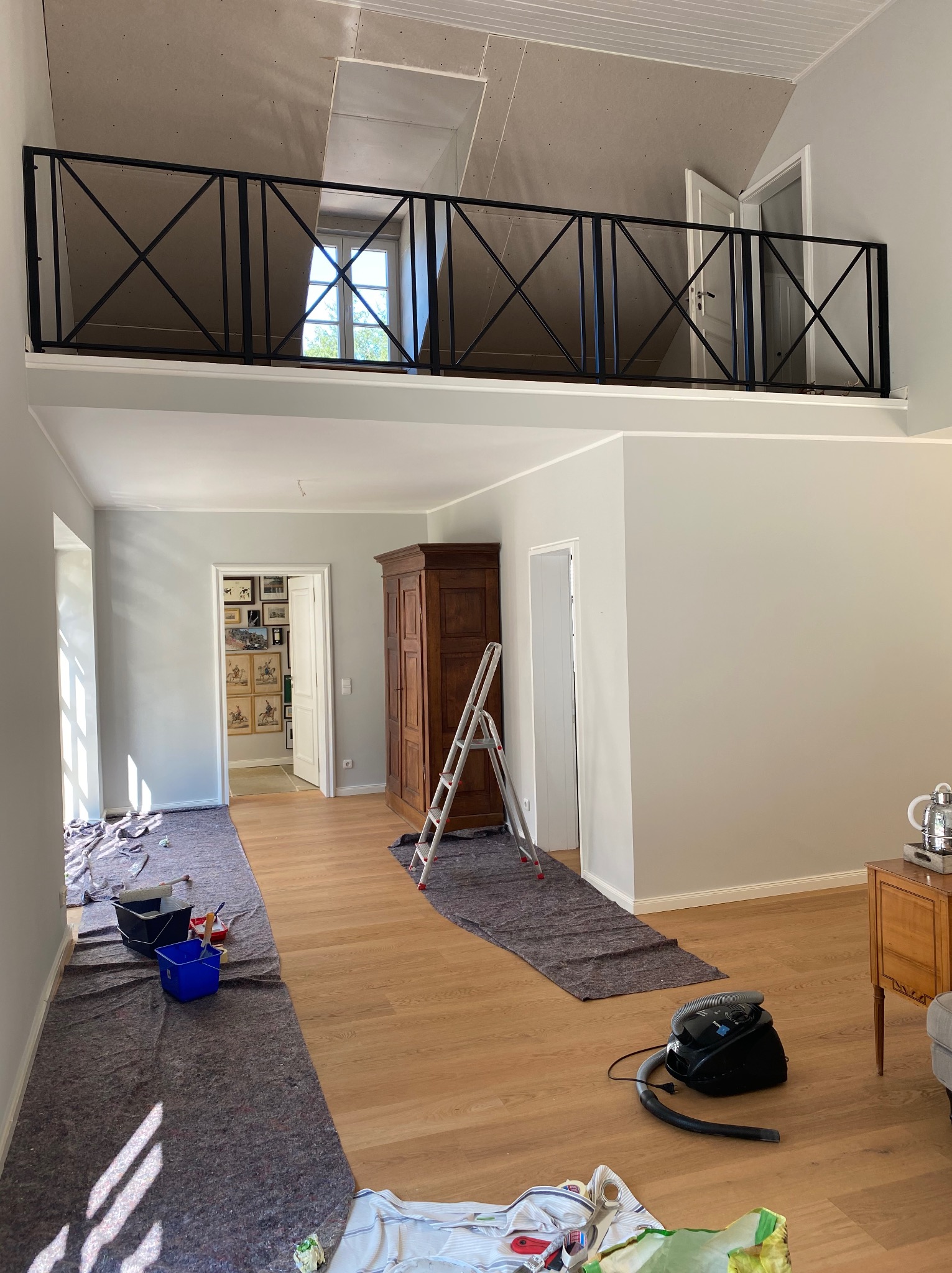 Dwupoziomowe wnętrze w trakcie malowania: zabezpieczona podłoga, drabina oparta o ścianę, widoczne narzędzia malarskie. Ściany w jasnym odcieniu szarości, sufit w trakcie prac.