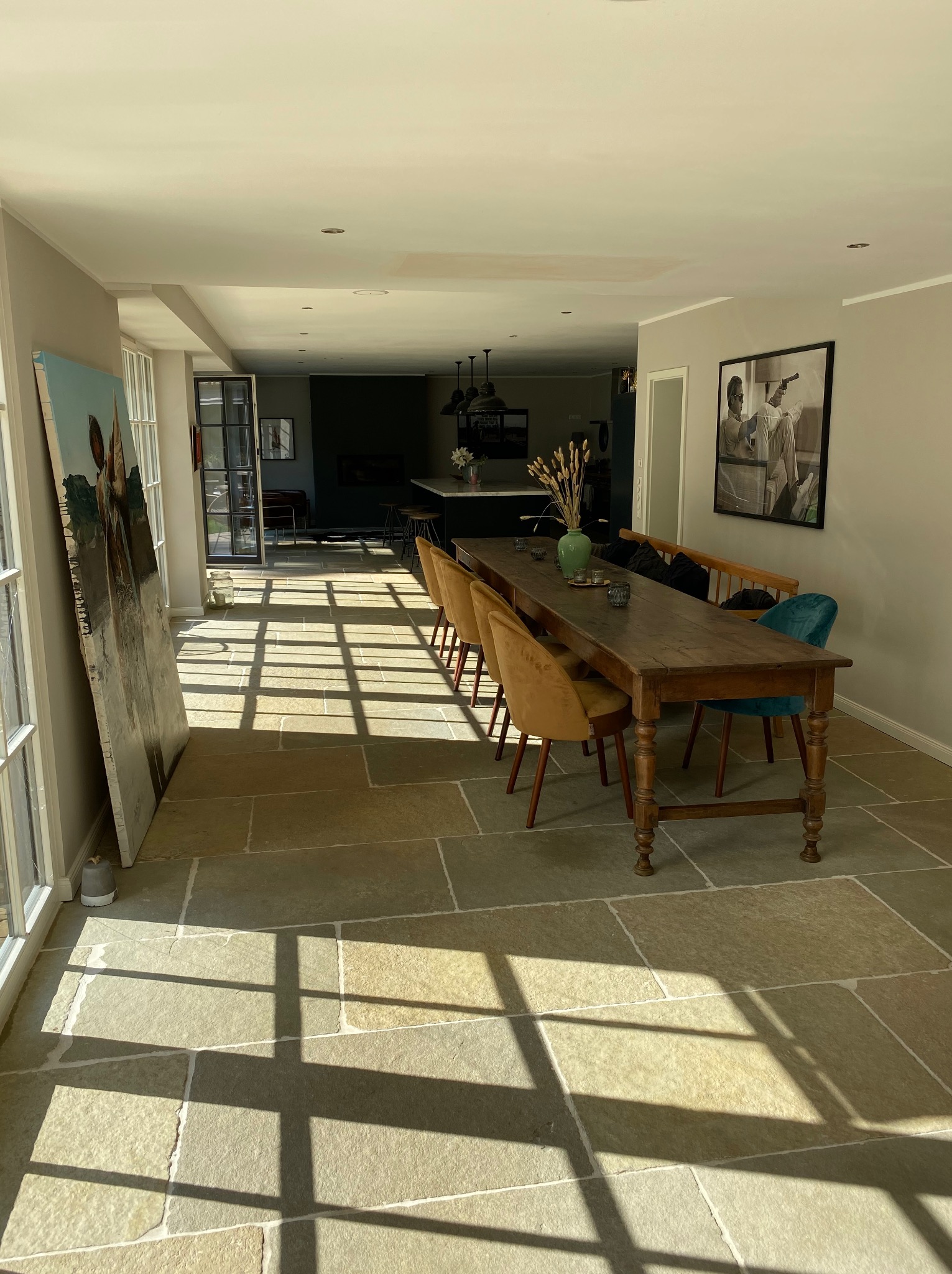 Jasne wnętrze z kamienną podłogą w odcieniach beżu, długim drewnianym stołem i krzesłami. W tle widoczna kuchnia i obraz oparty o ścianę.