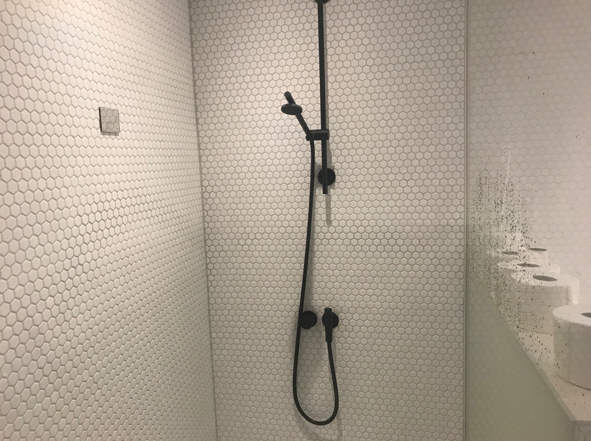 Biała łazienka z heksagonalnymi płytkami i czarną armaturą natryskową. Widoczny fragment szklanej kabiny z zaciekami i półka z rolkami papieru toaletowego.